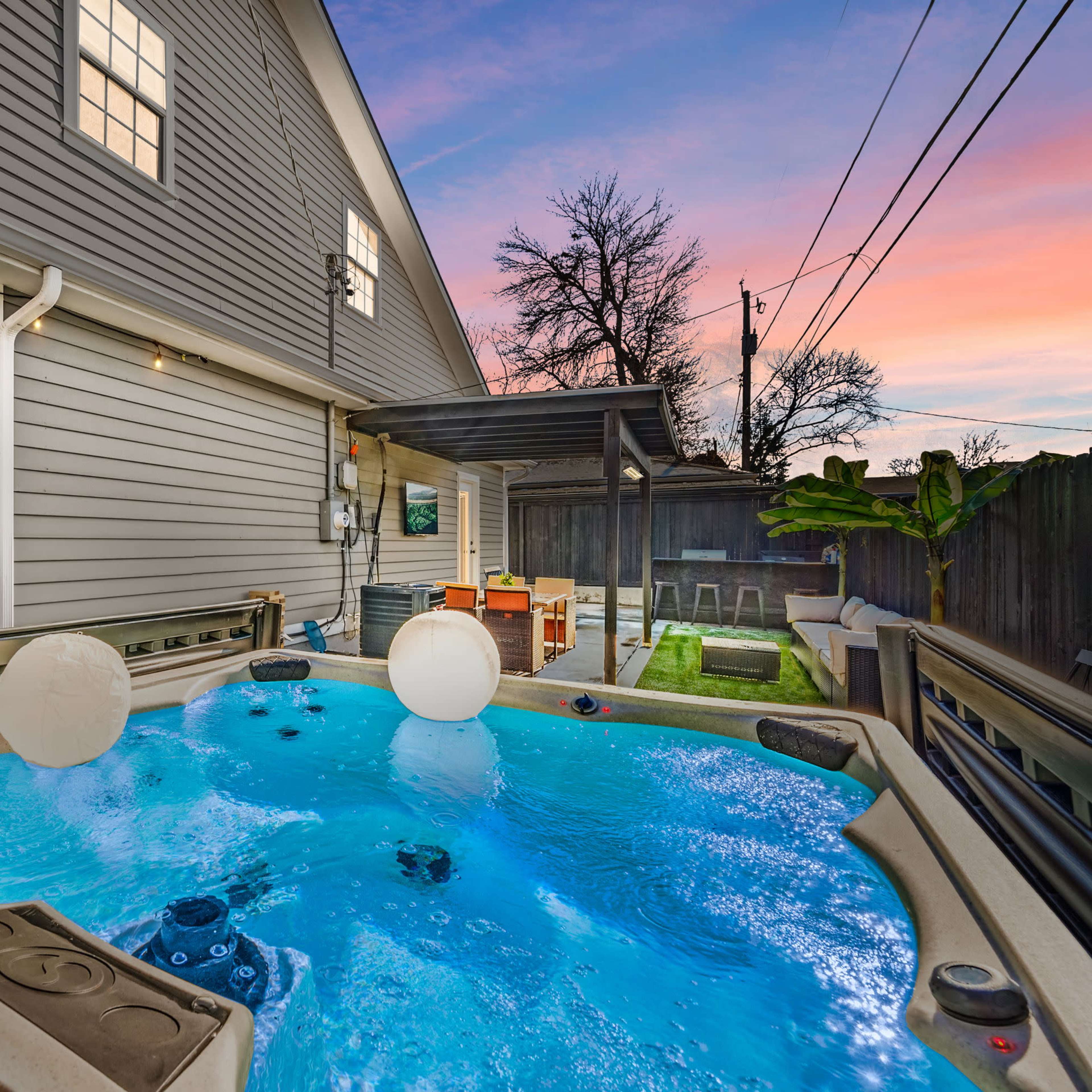 A backyard scene features a hot tub with two floating balls, surrounded by a house and patio area furnished with a dining set.
