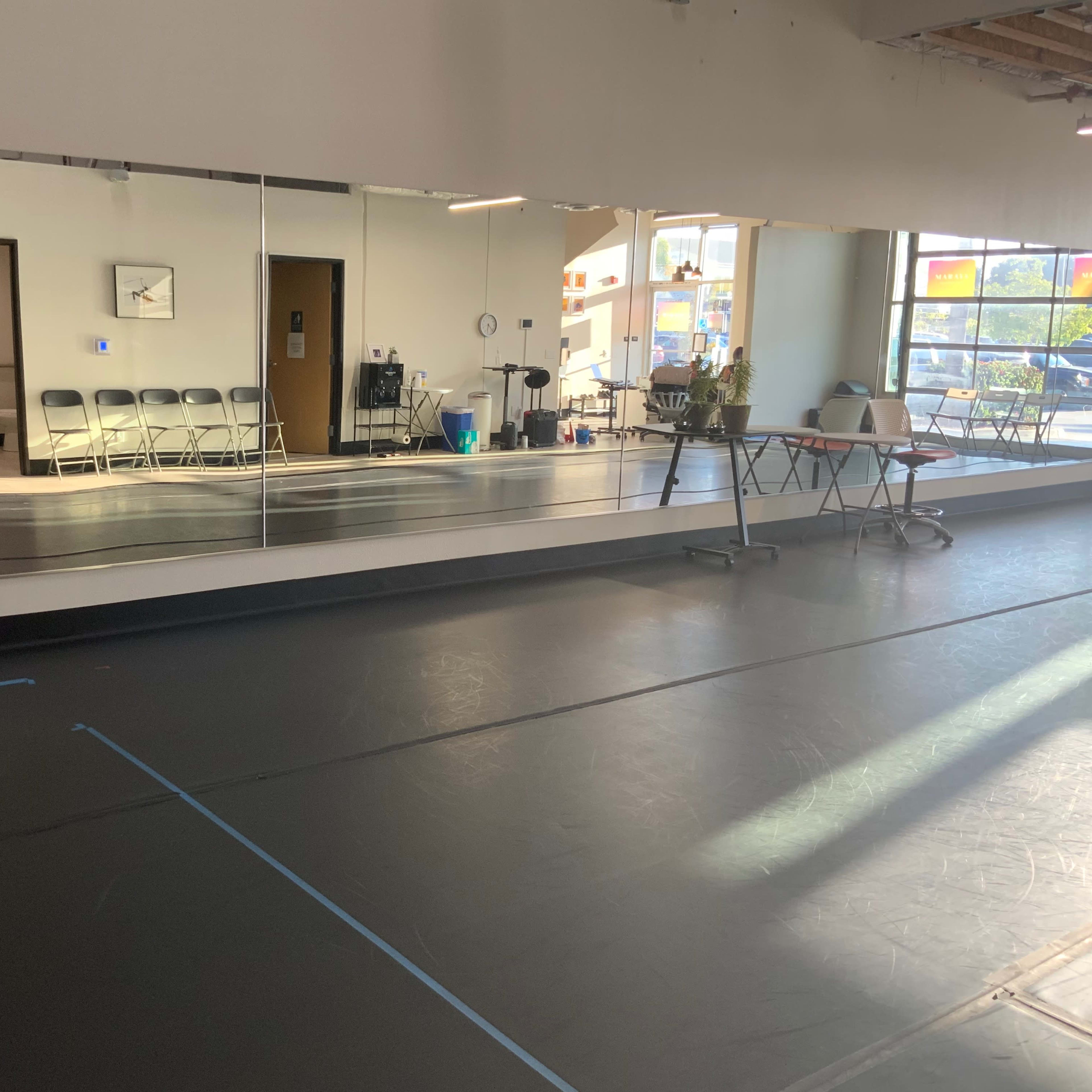 A spacious dance studio with mirrors along one wall, a smooth black floor, and several folding chairs arranged in the background.
