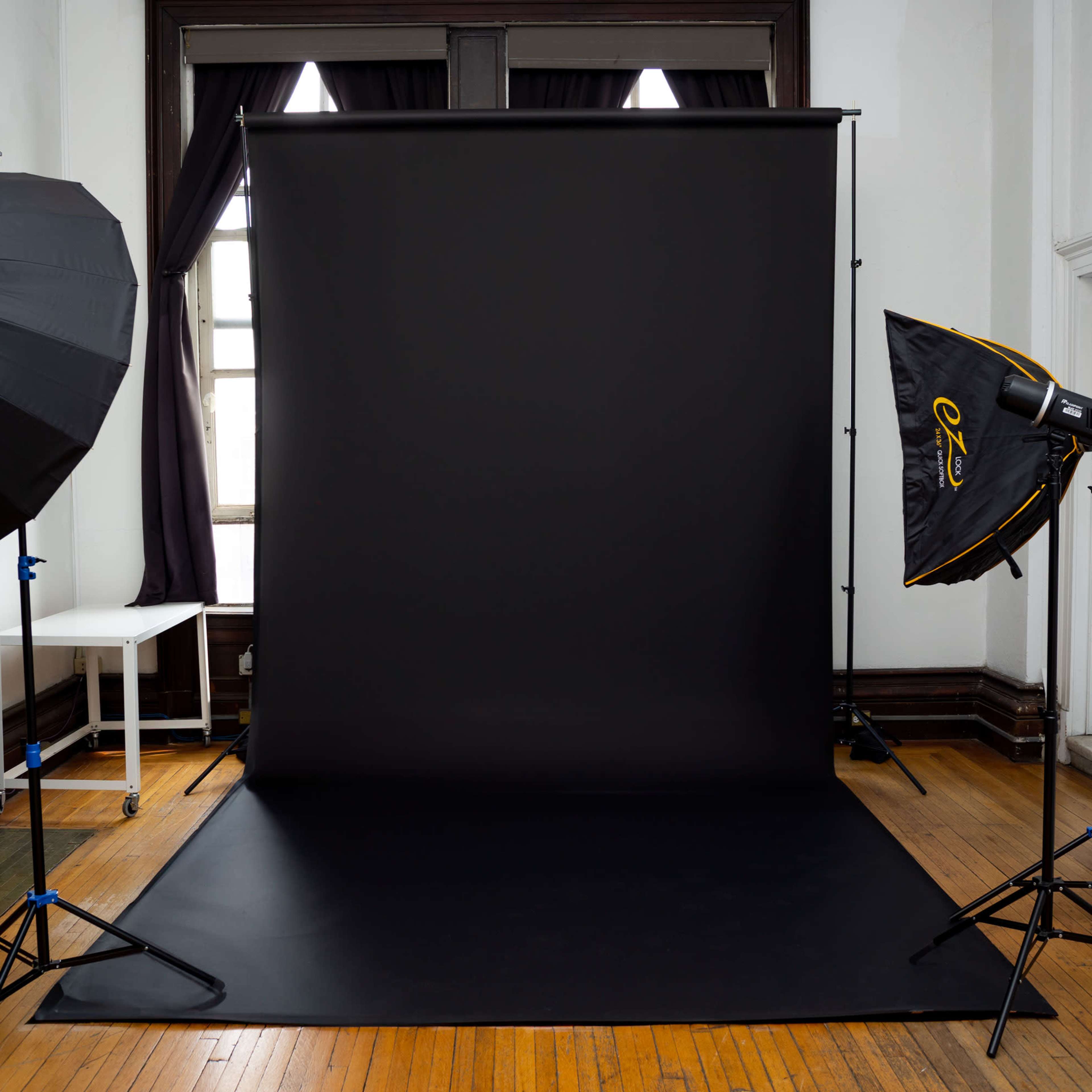 A photography studio setup with a black backdrop, softbox lights on either side, and wooden flooring.