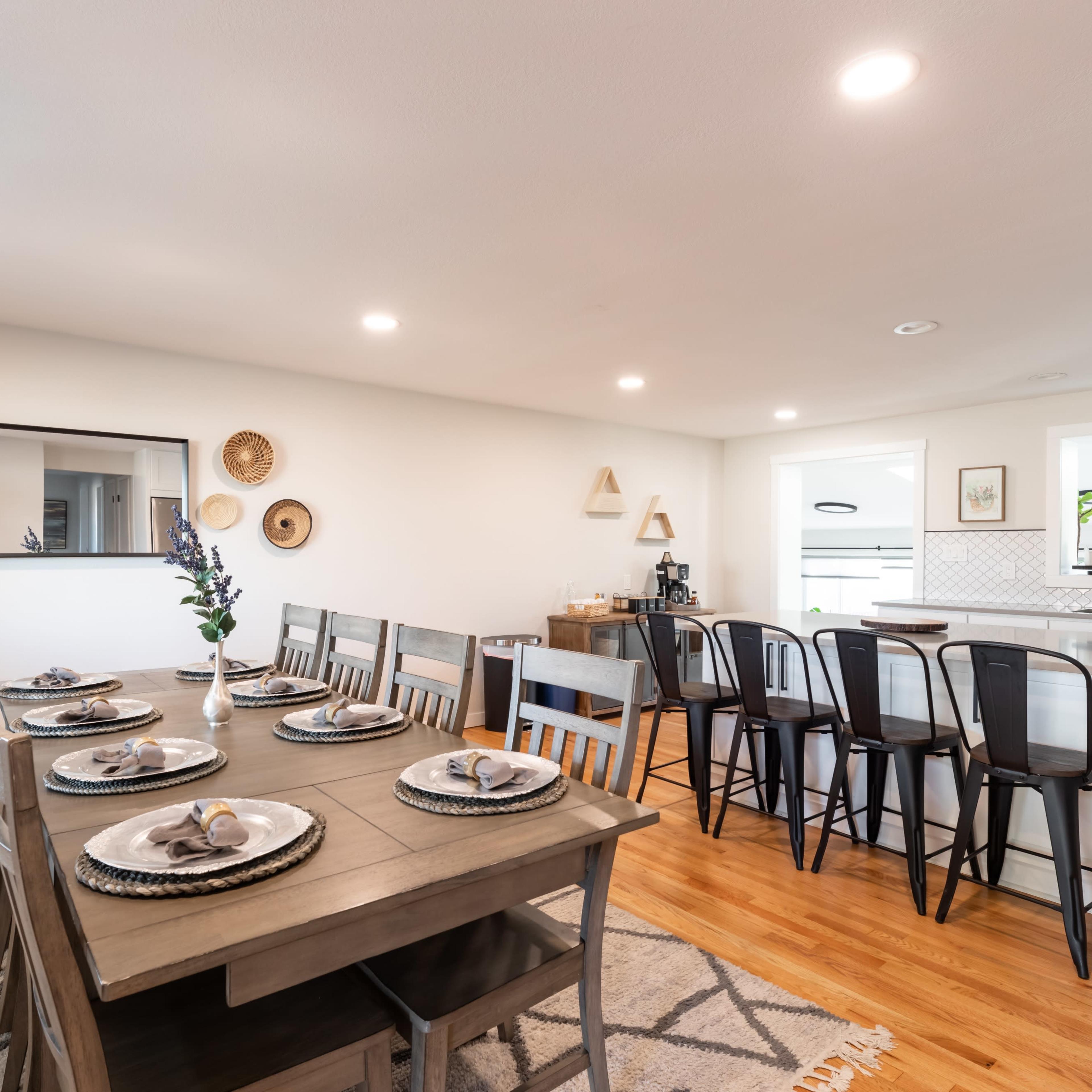 A dining area features a table set for eight with plates and utensils, adjacent to a kitchen with a breakfast bar and various decor on the walls.