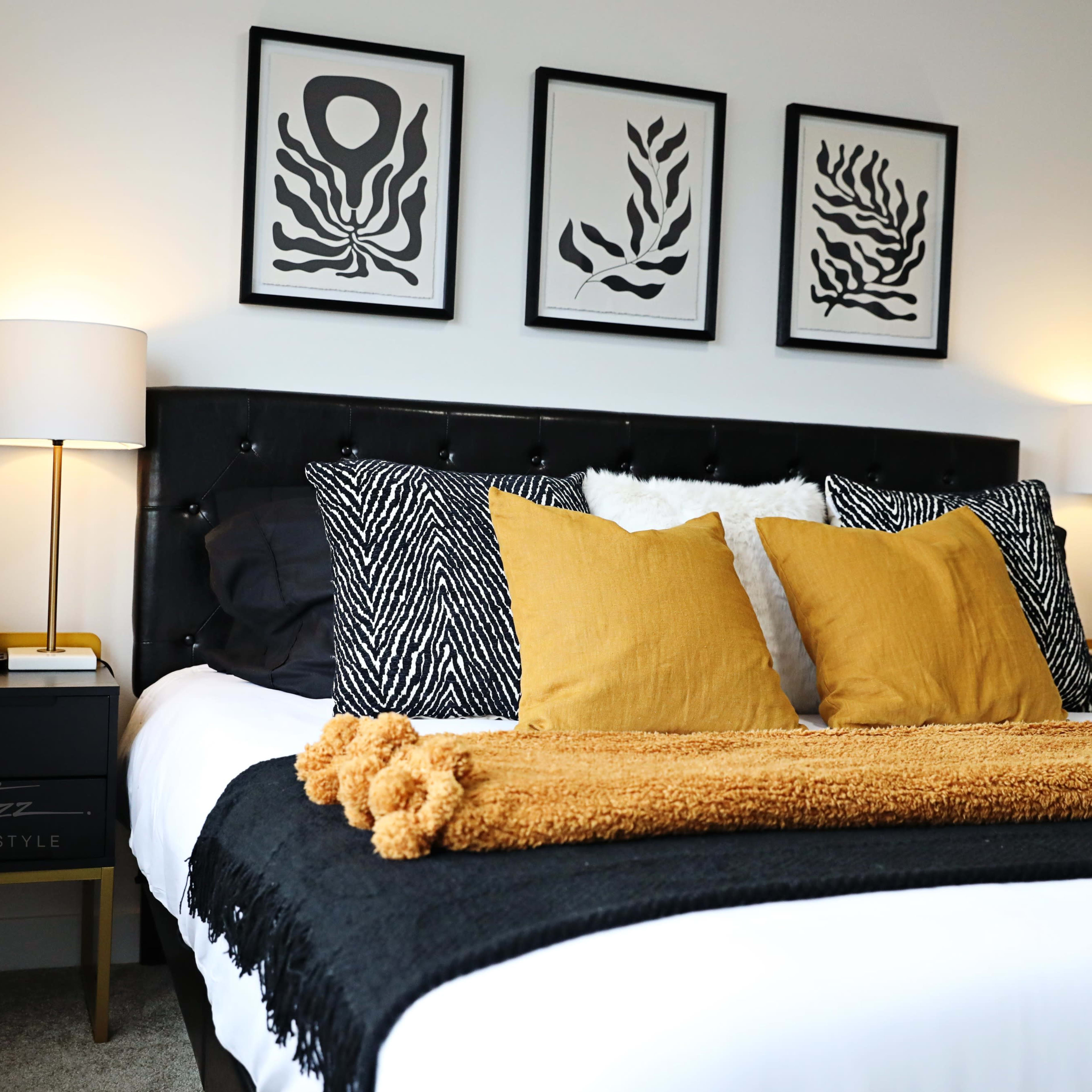 A modern bedroom featuring a black upholstered bed with decorative pillows and a mustard-colored throw, flanked by bedside tables with lamps and framed wall art.