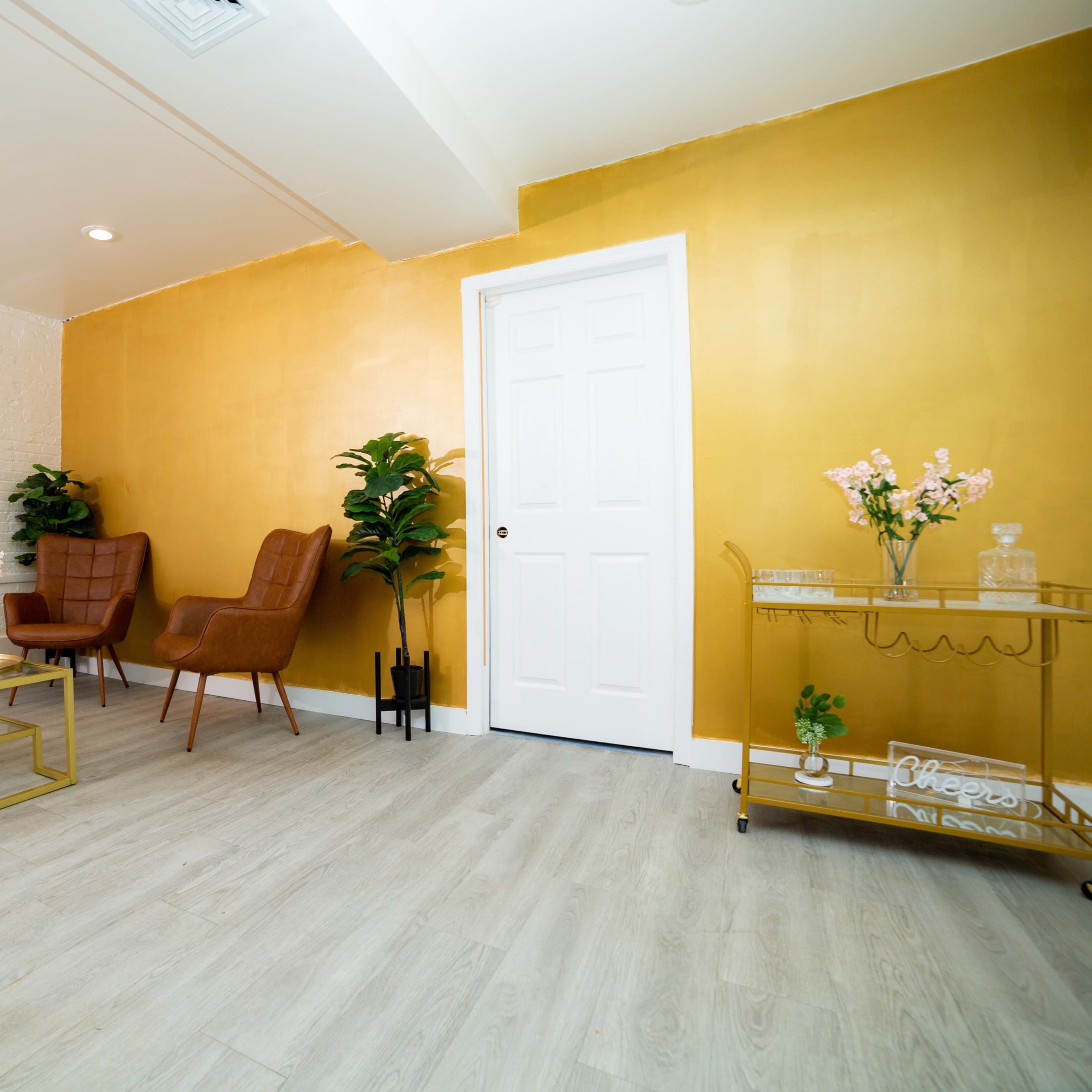 A room with a golden yellow accent wall, two brown armchairs, a small coffee table, and a decorative cart with plants and flowers.