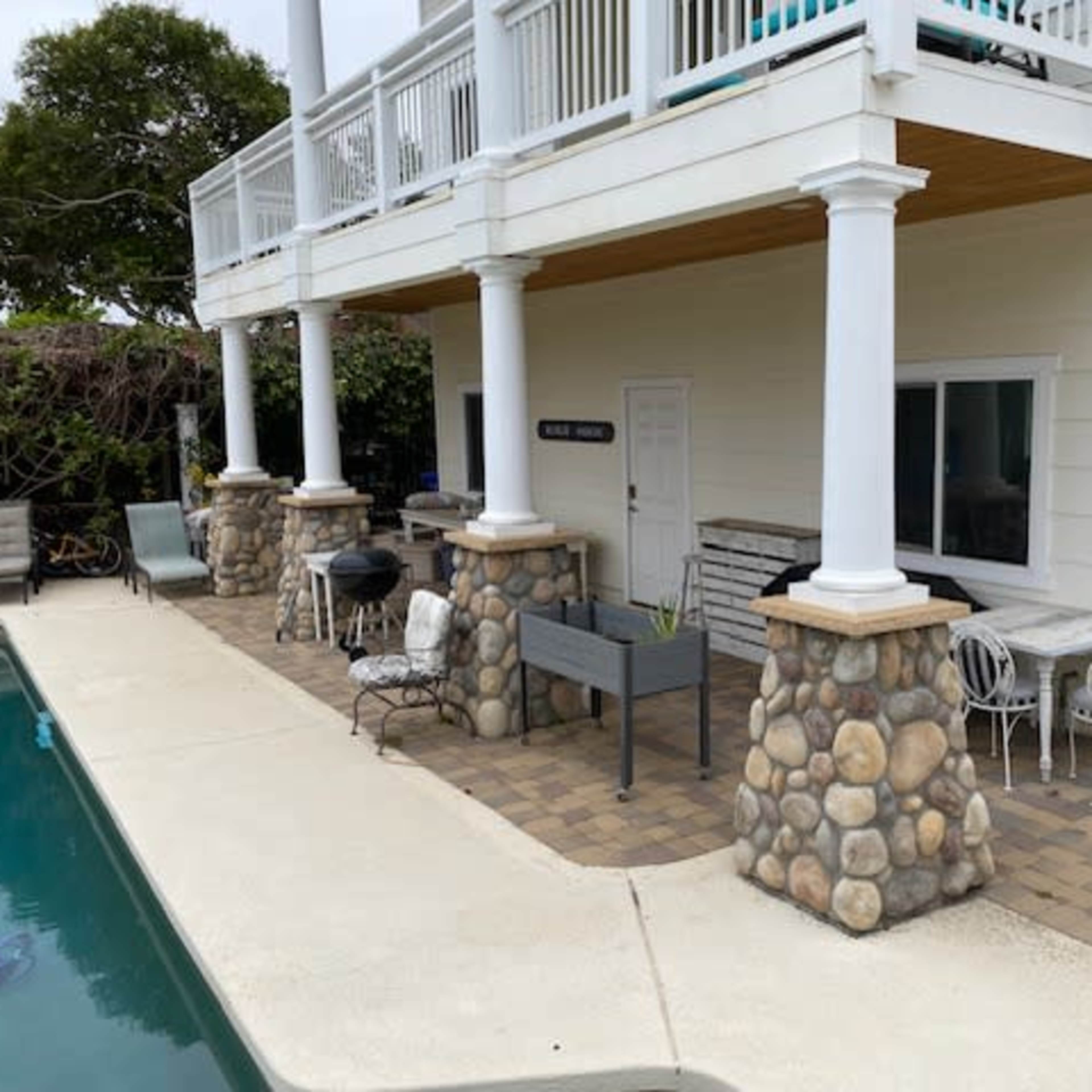 A patio area beside a swimming pool, featuring a house with a light-colored exterior, stone column bases, and some outdoor furniture.