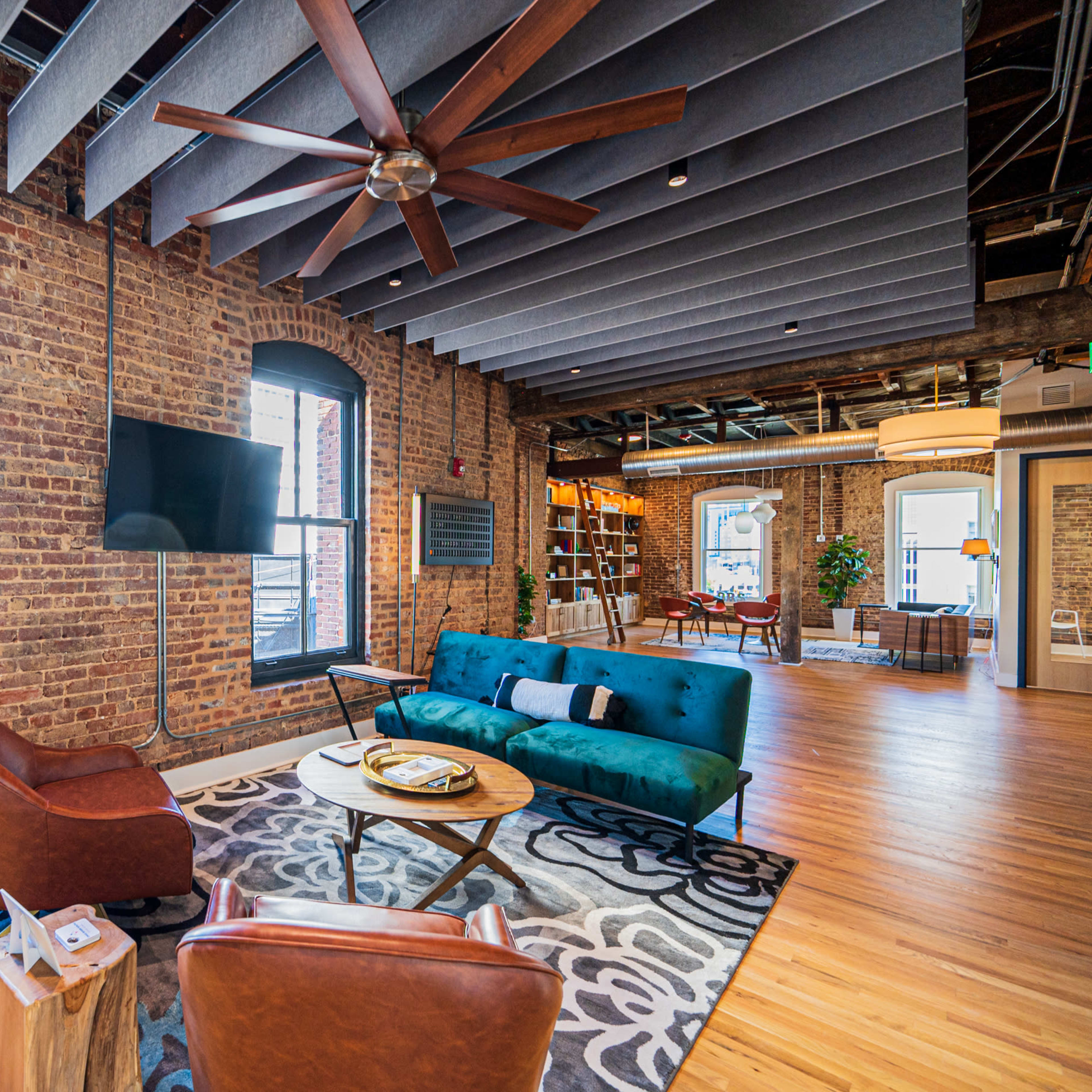 A room featuring exposed brick walls, a blue sofa, two brown leather chairs, a round coffee table, and a ceiling fan, with large windows allowing natural light to enter.