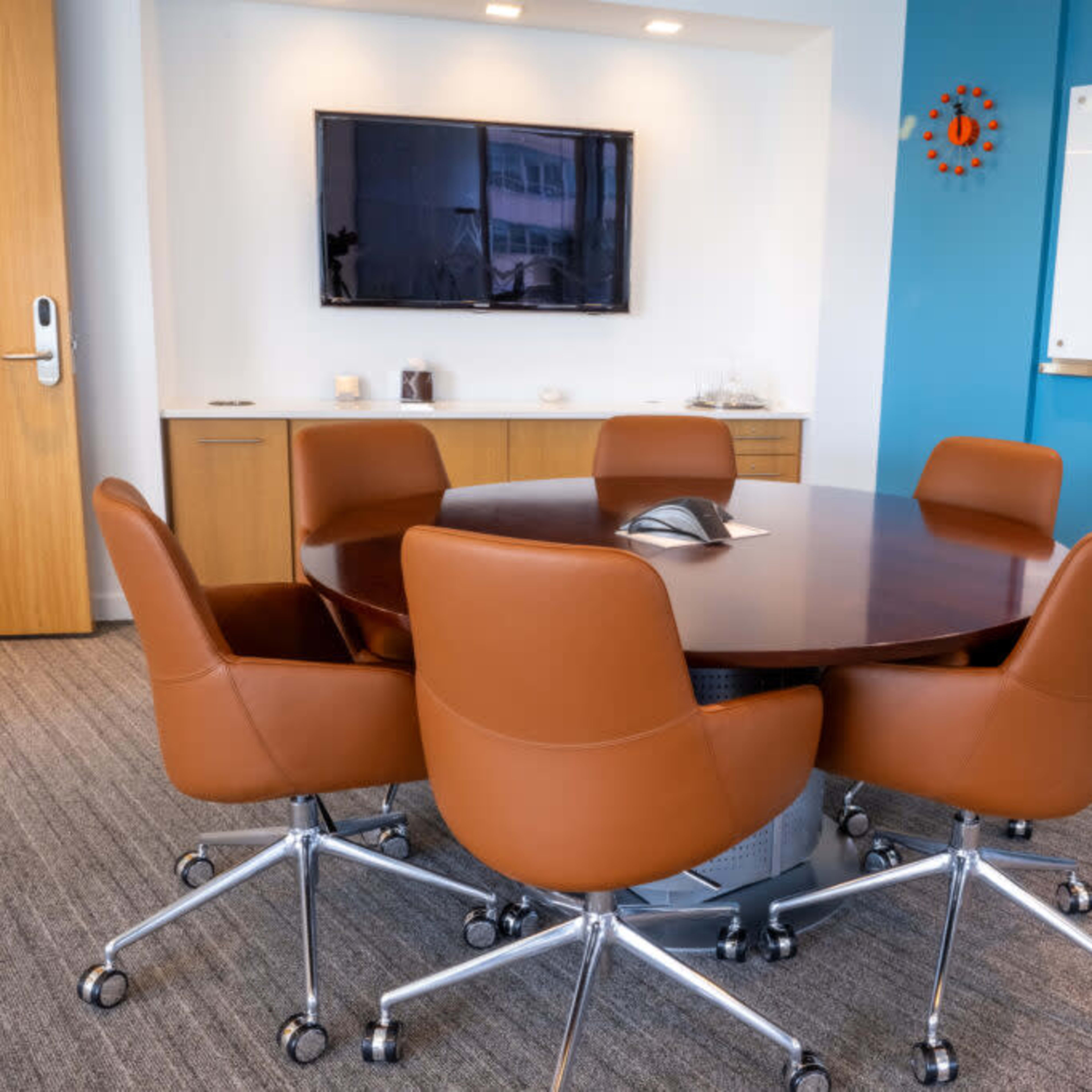 A modern conference room features a round wooden table surrounded by brown leather chairs and a wall-mounted television.