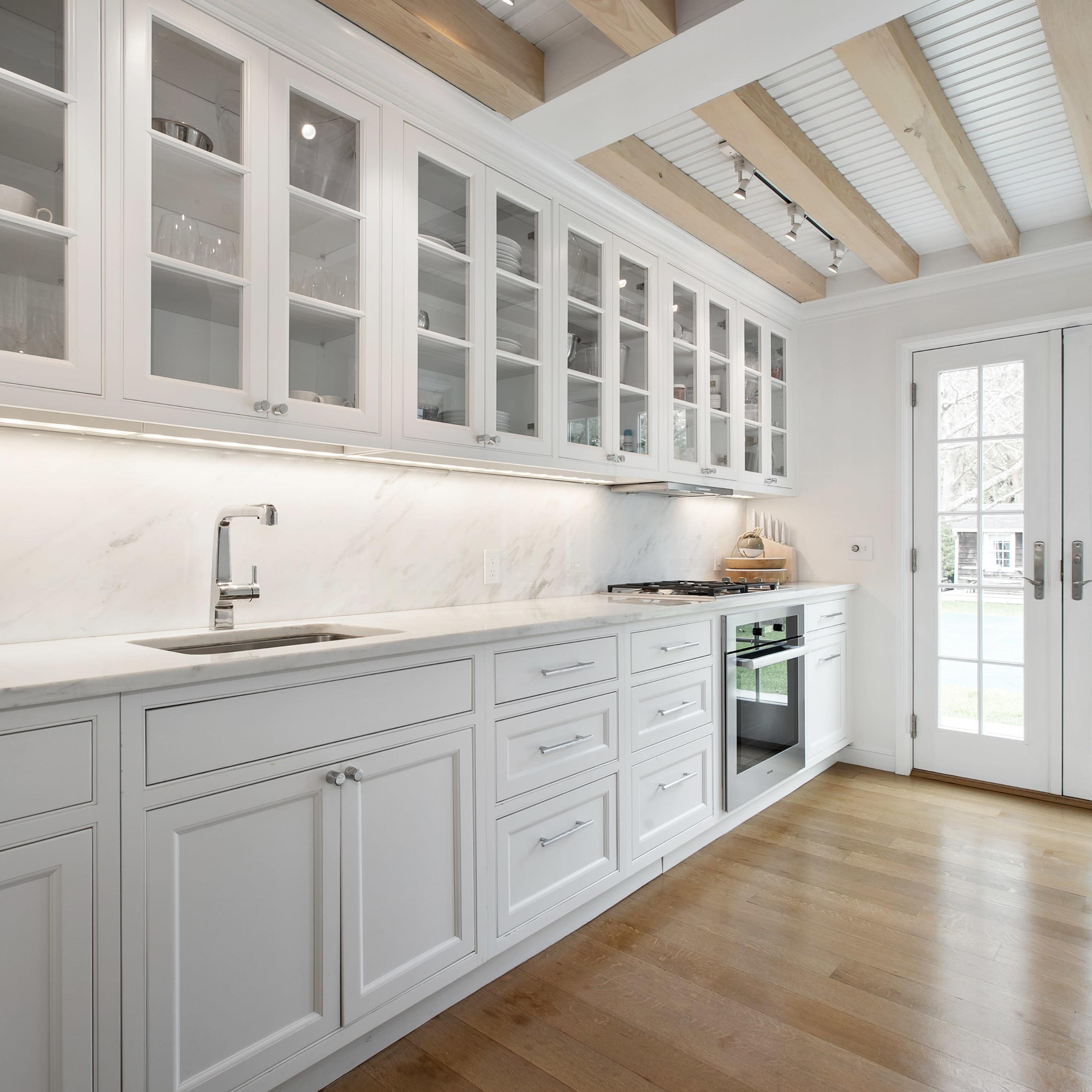 The kitchen features white cabinetry, glass-fronted cabinets, a marble countertop, and a large window with double doors leading to the outdoors.