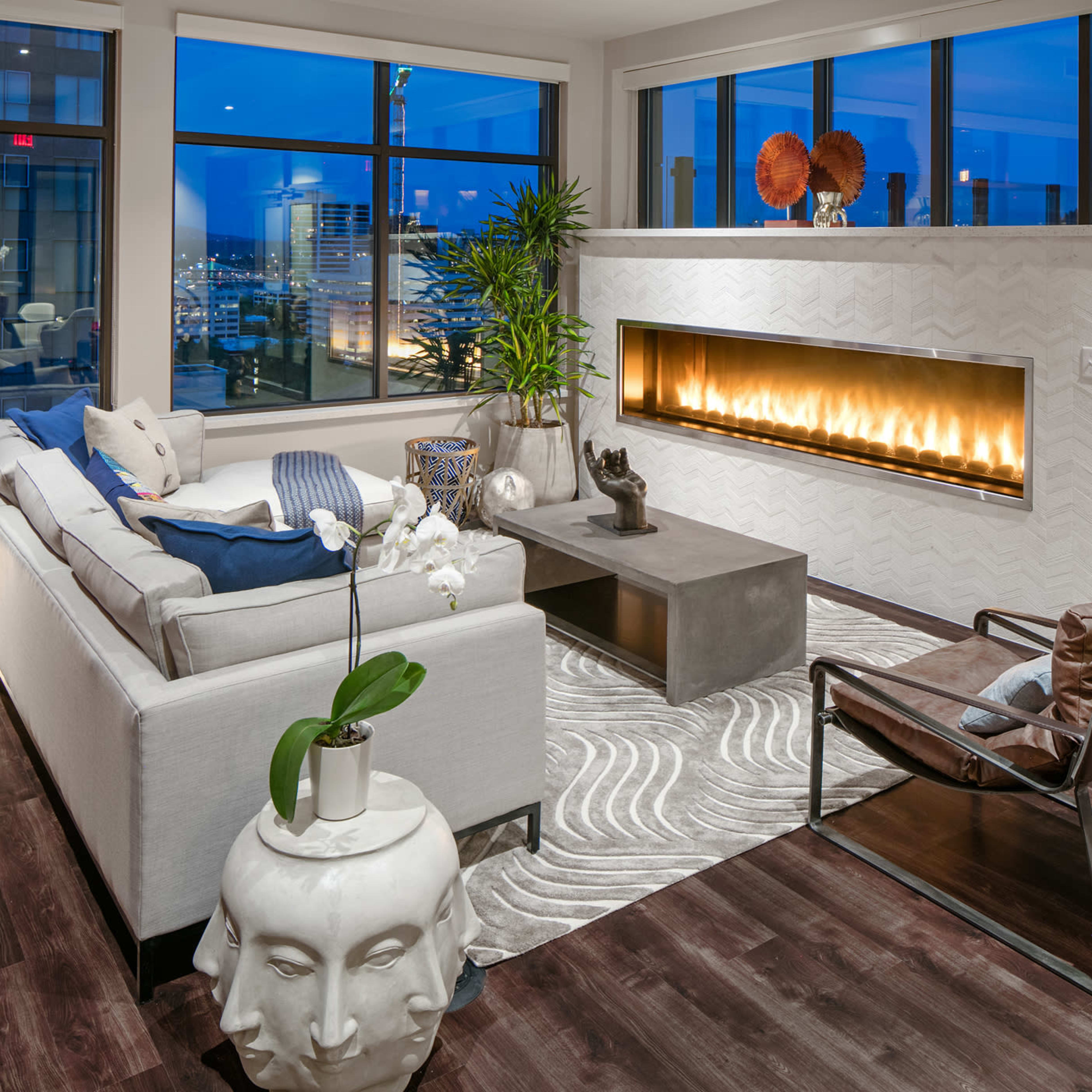 A modern living room features a sleek fireplace, large windows with city views, and stylish furniture arranged on a patterned rug.