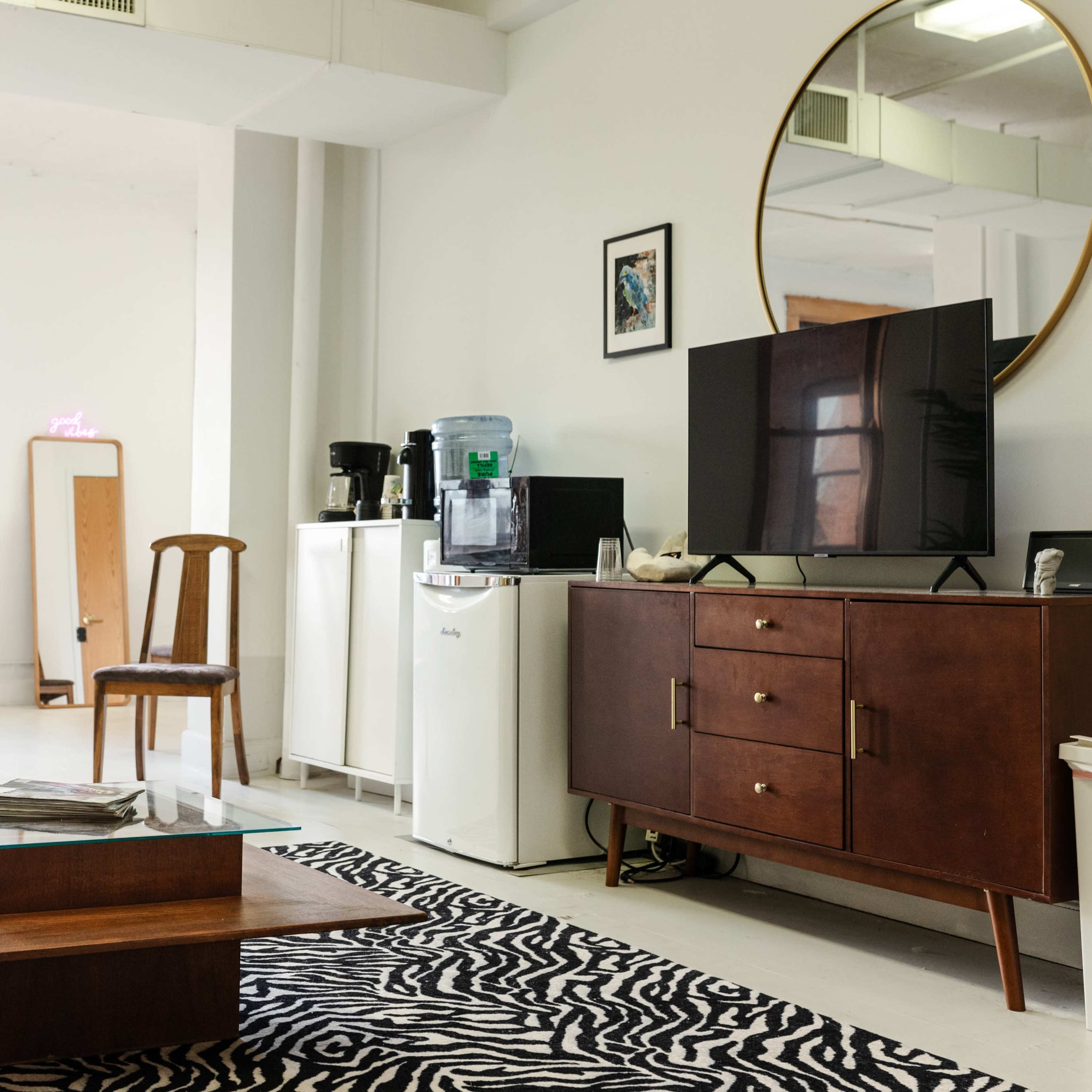 A modern, furnished living room with a large mirror, a television on a wooden cabinet, a coffee maker, and a table on a zebra-striped rug.
