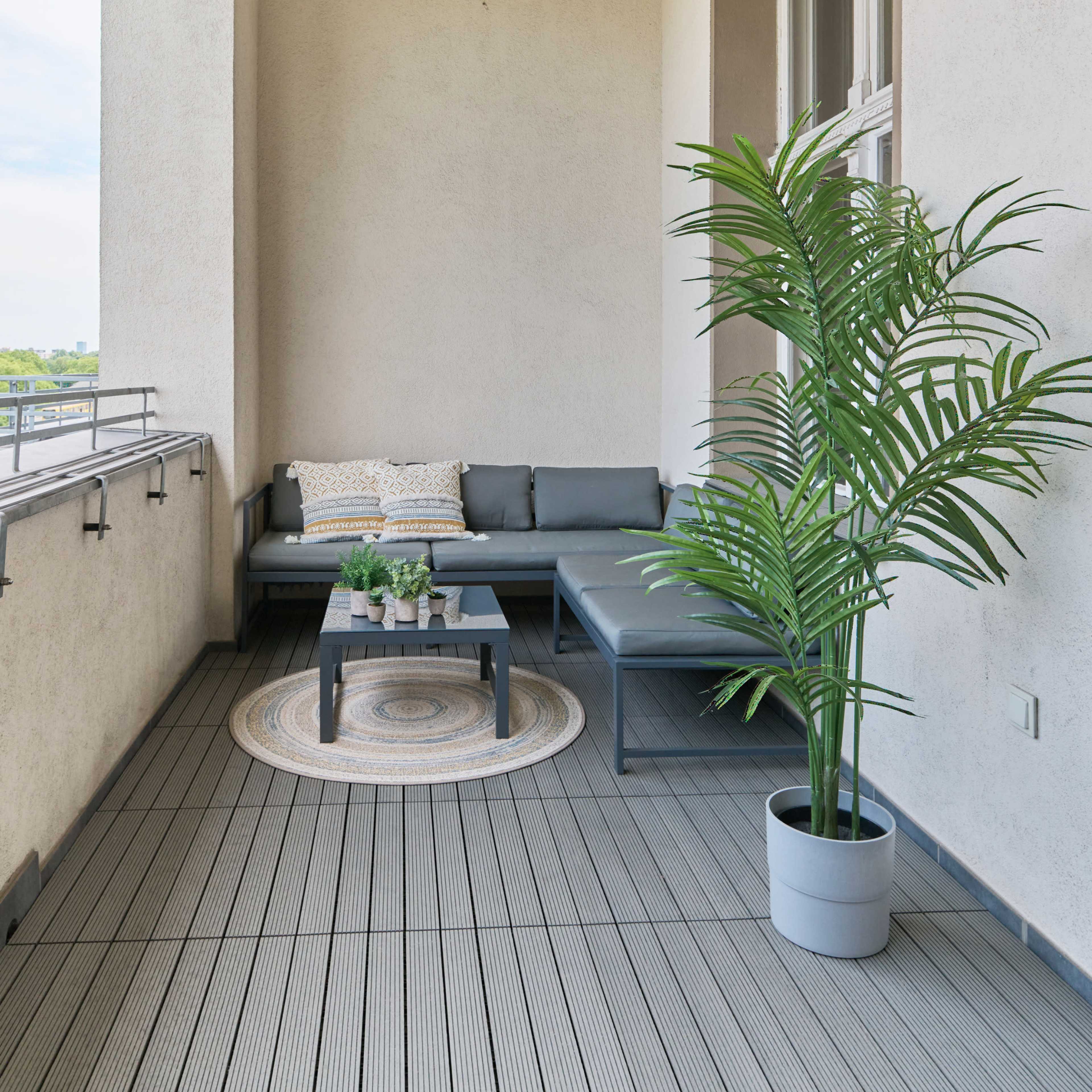 A balcony featuring a gray sectional sofa, a small black coffee table, a round rug, a potted plant, and a wall-mounted light fixture.