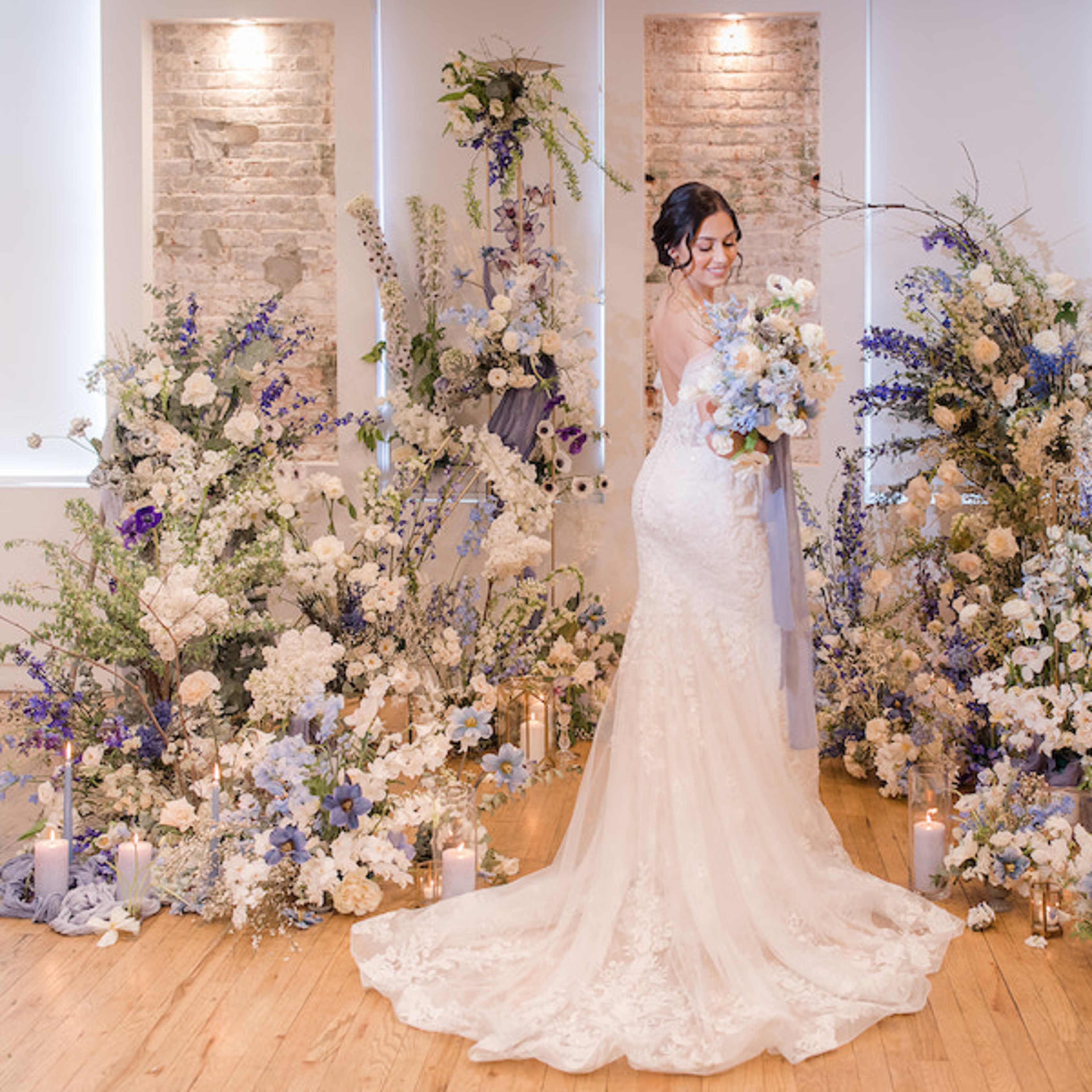 A bride in a fitted lace gown stands amidst elegant floral arrangements featuring white and blue blooms, with candles arranged around them.