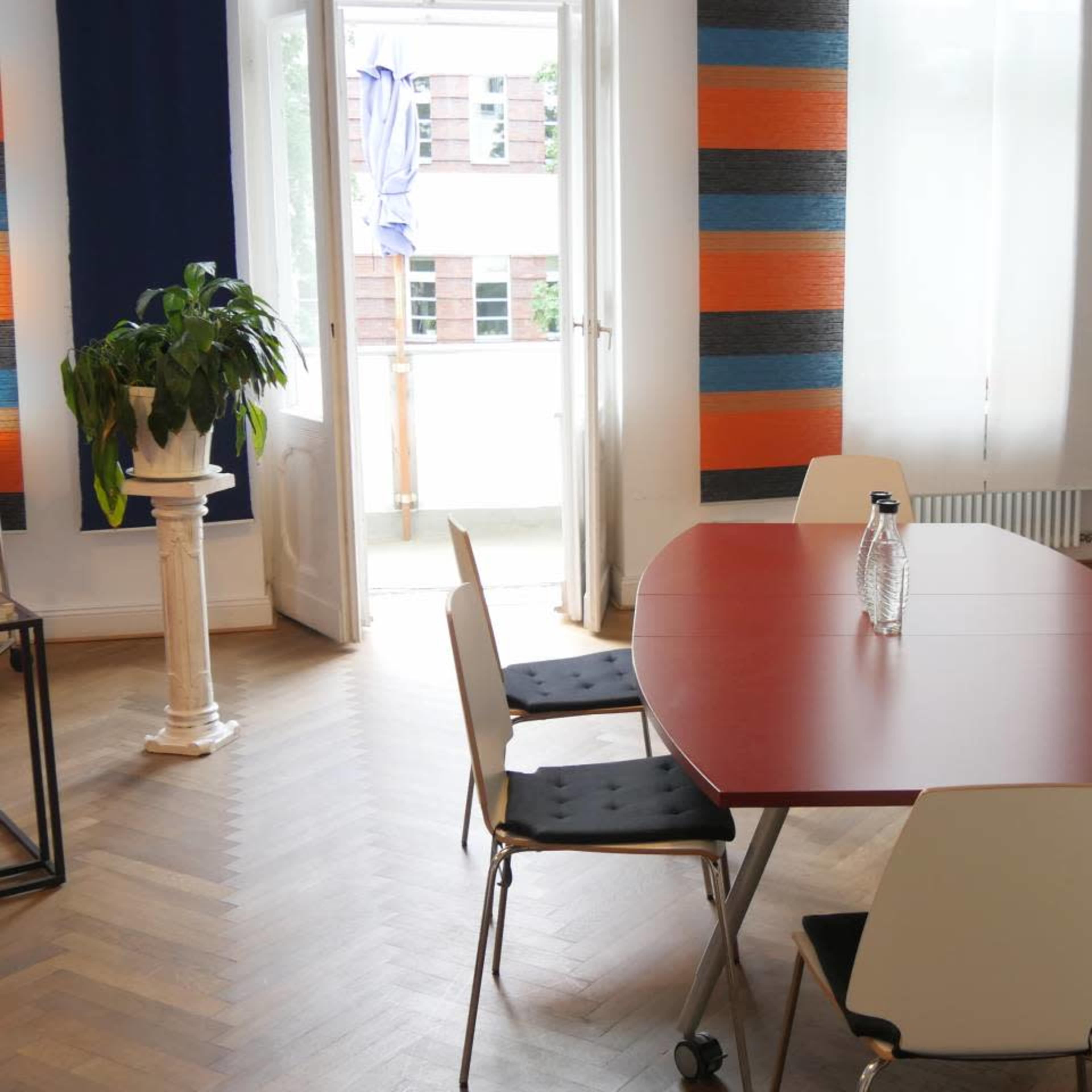 A modern meeting room with a red oval table surrounded by five chairs, featuring large windows and a potted plant.