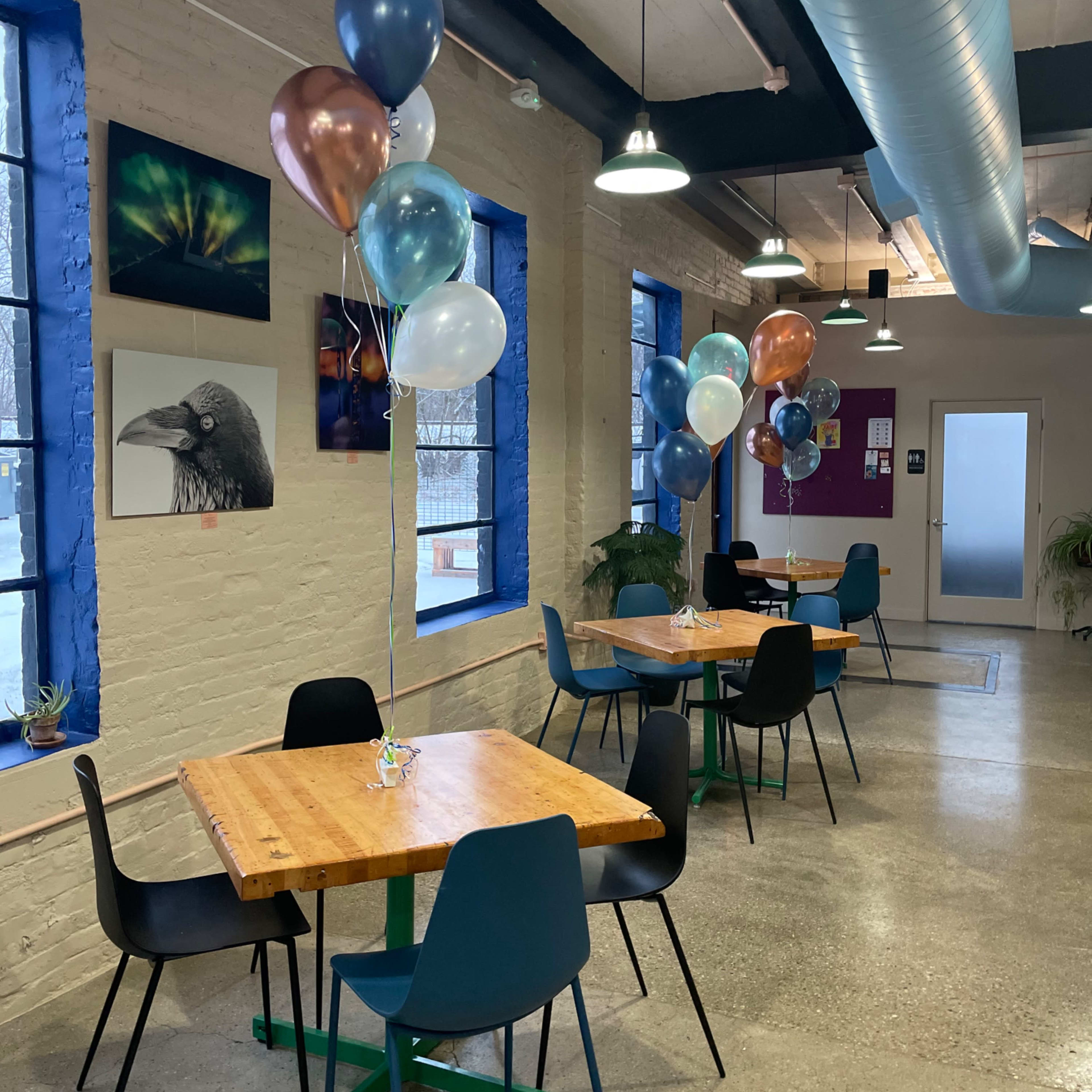 A modern café features several wooden tables surrounded by chairs and is decorated with clusters of balloons in various colors.