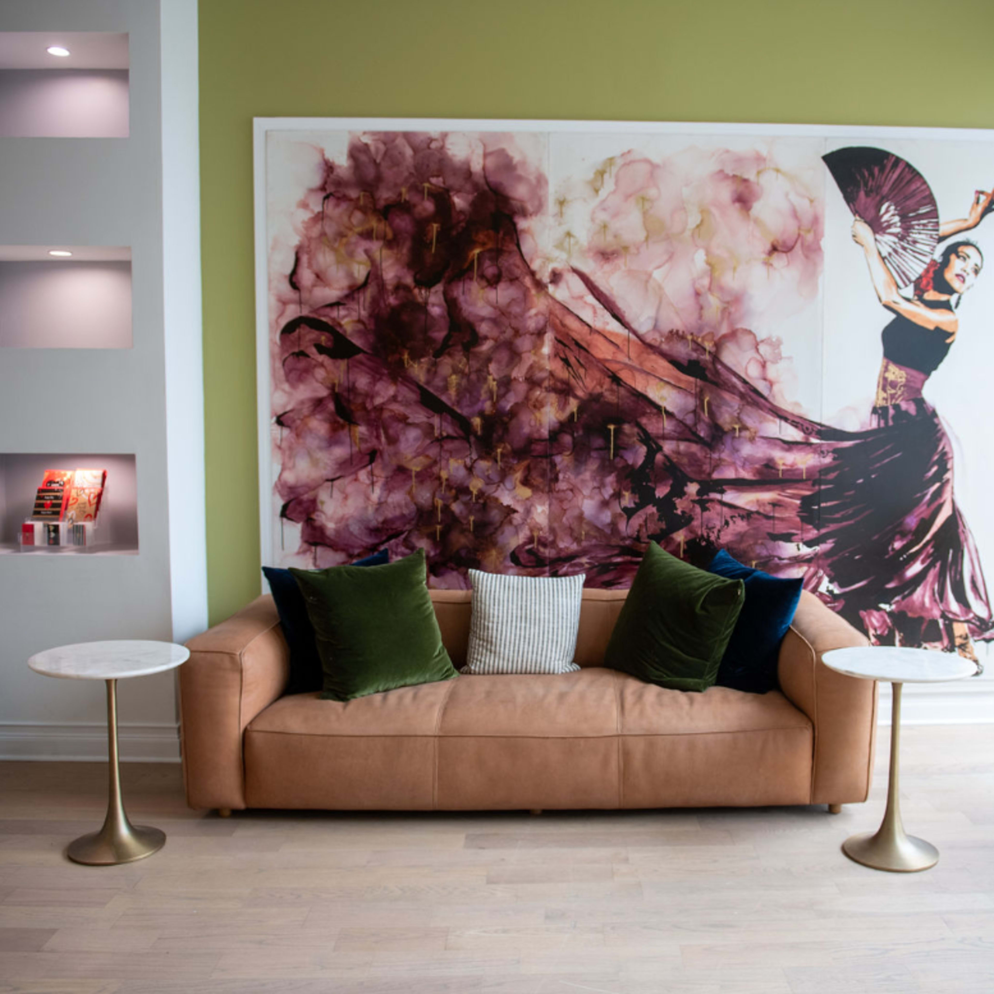 A modern seating area featuring a brown sofa with green and white cushions, flanked by small round tables and a large wall mural depicting a dancer.