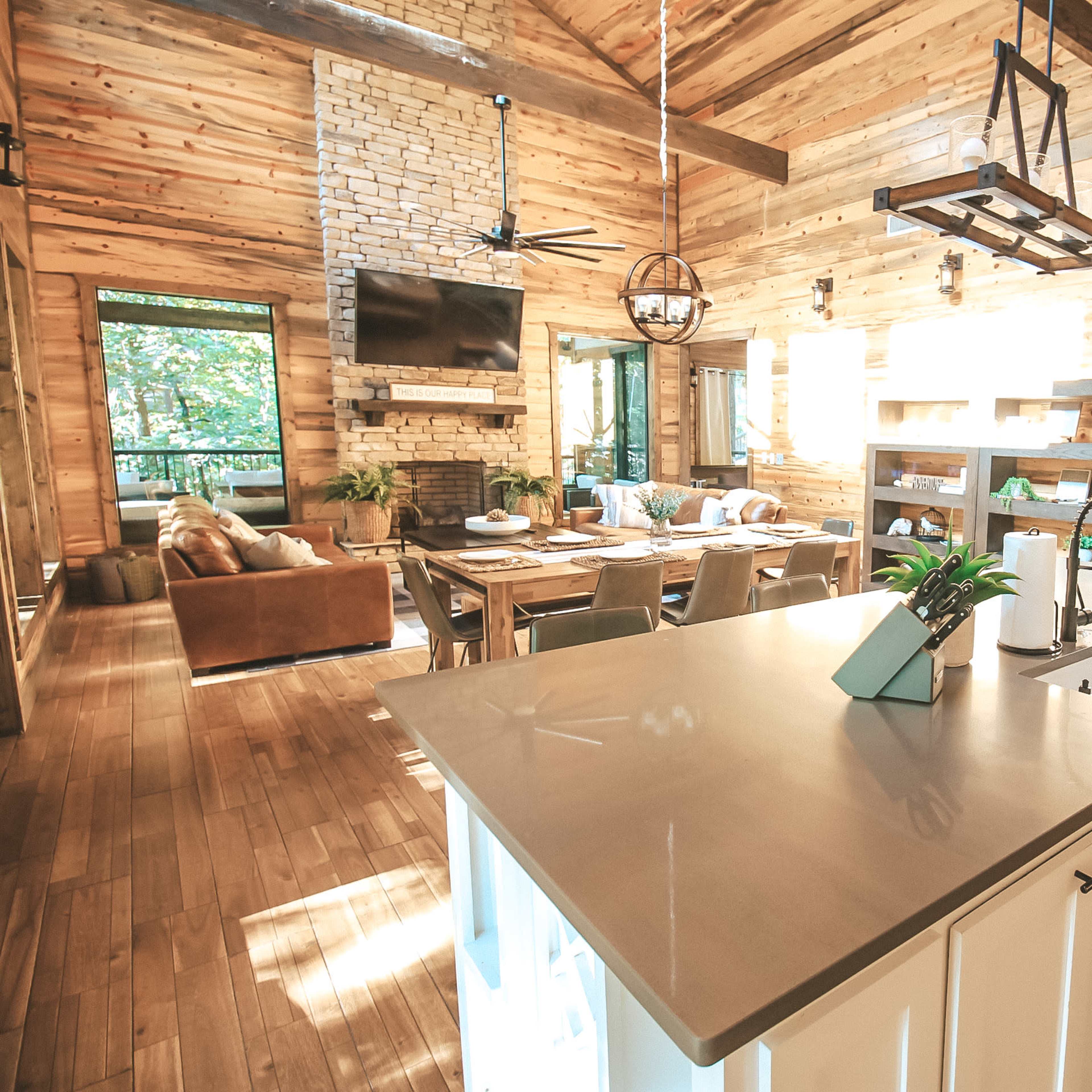 A spacious wood-paneled cabin interior with an open kitchen, dining area, and living space featuring a stone fireplace and large windows.