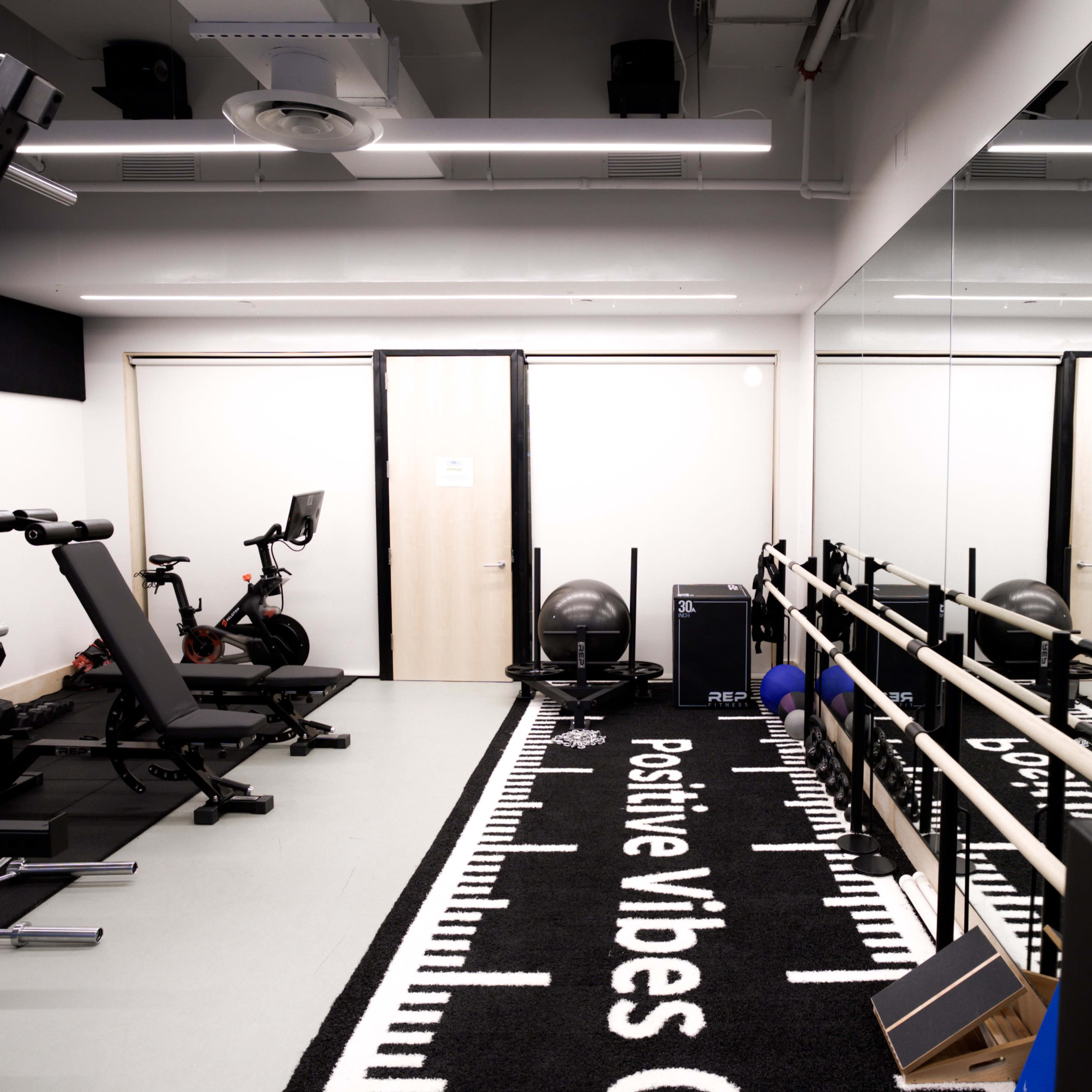 A modern gym space equipped with weights, exercise machines, a stability ball, and mirrors, featuring a carpet with the phrase "Positive Vibes Only."