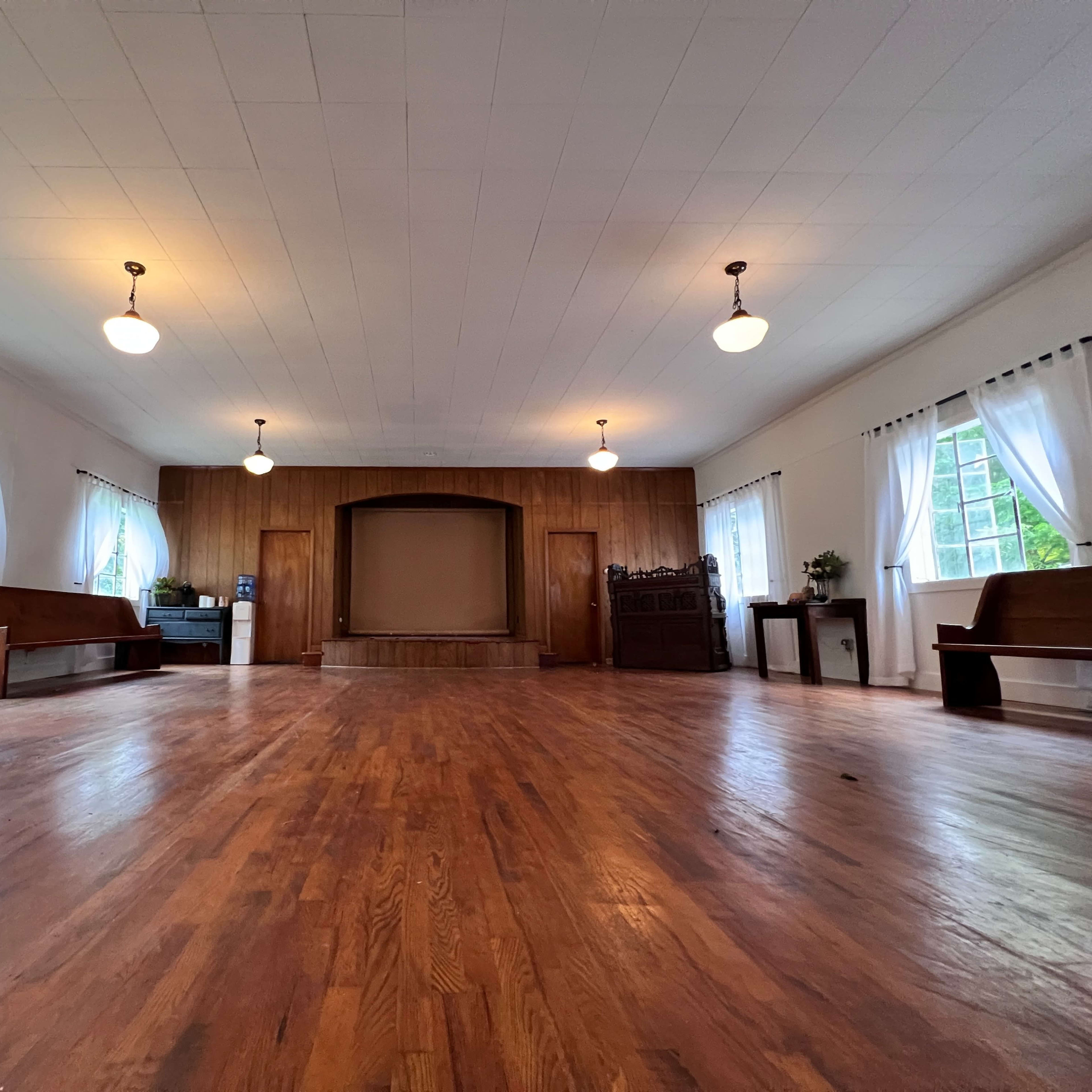 A spacious, empty hall with wooden floors, benches along the sides, and large windows with white curtains.