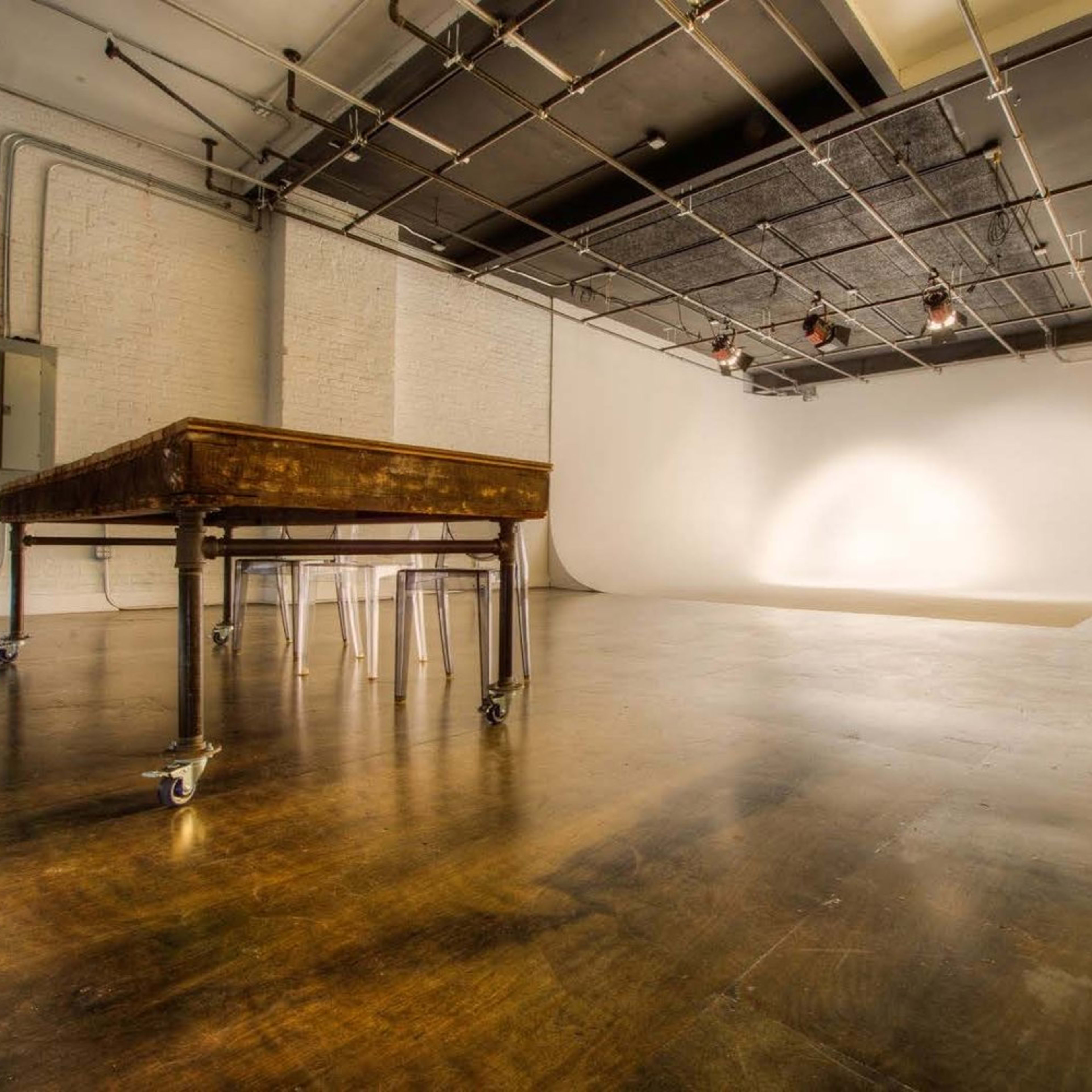 A spacious, minimalist studio features a large table on wheels and clear chairs, with a blank backdrop and overhead lighting.