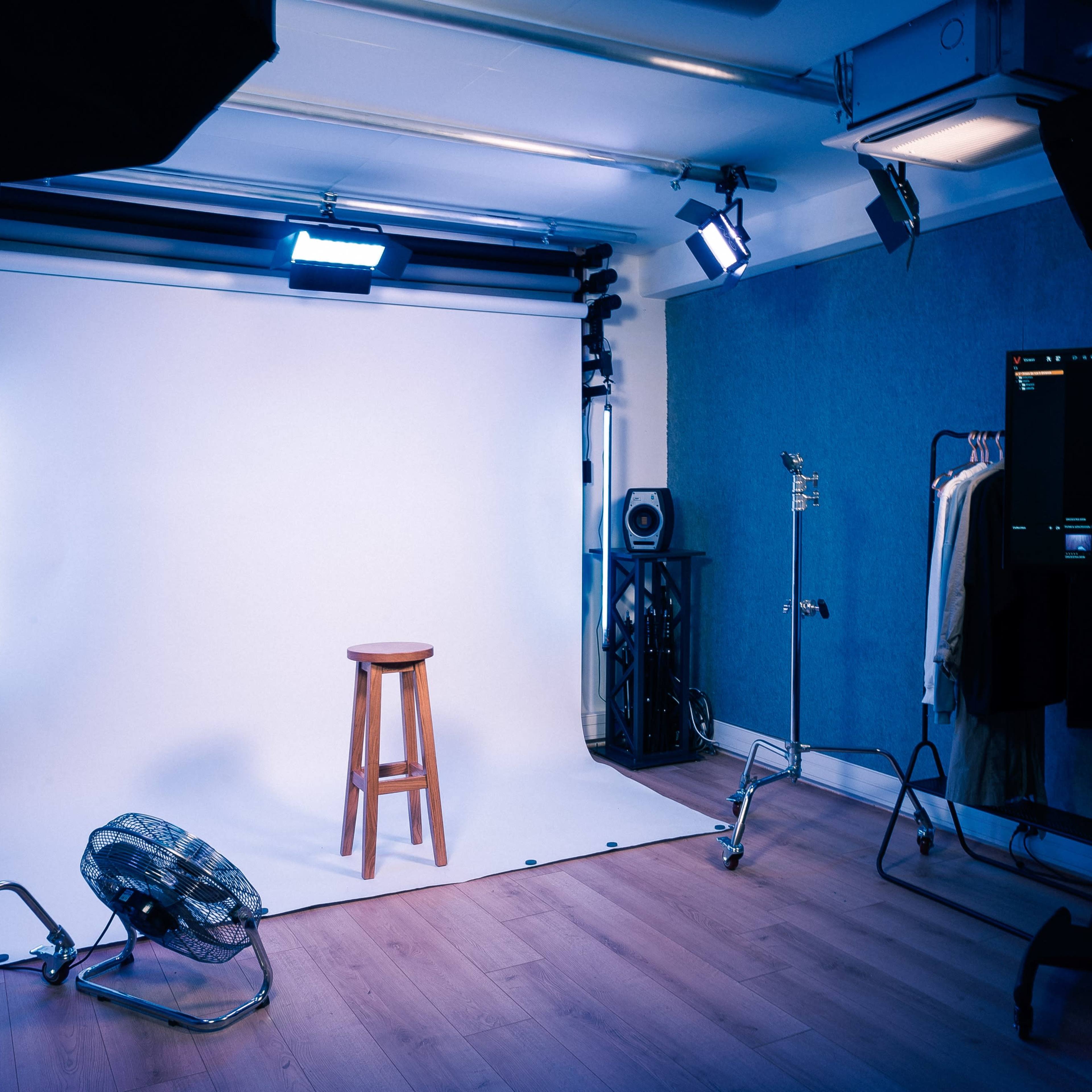 A photography studio with a wooden stool positioned in front of a white backdrop, surrounded by studio lights and various equipment.
