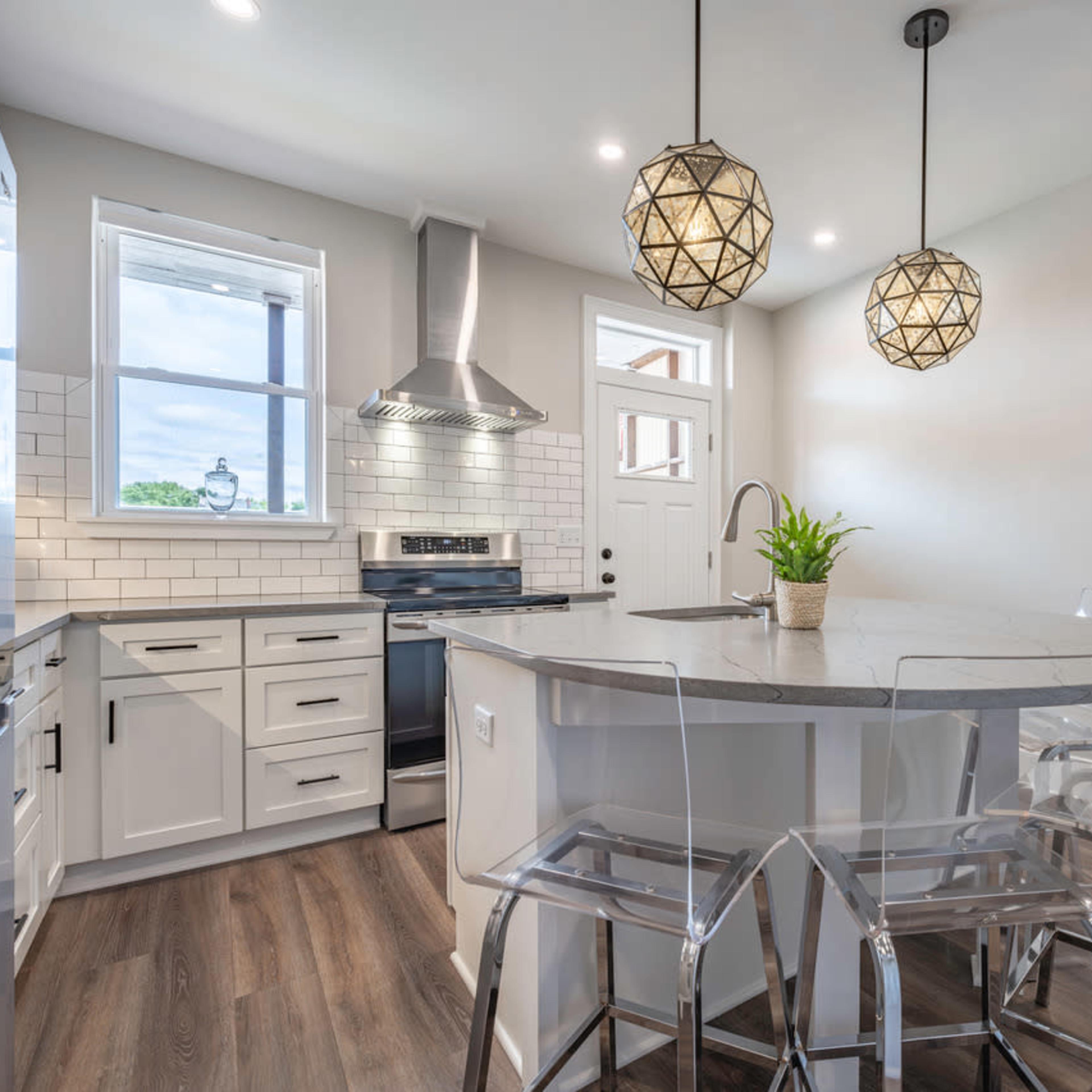 A modern kitchen features a round island with transparent stools, pendant lighting, stainless steel appliances, and a window above the sink.