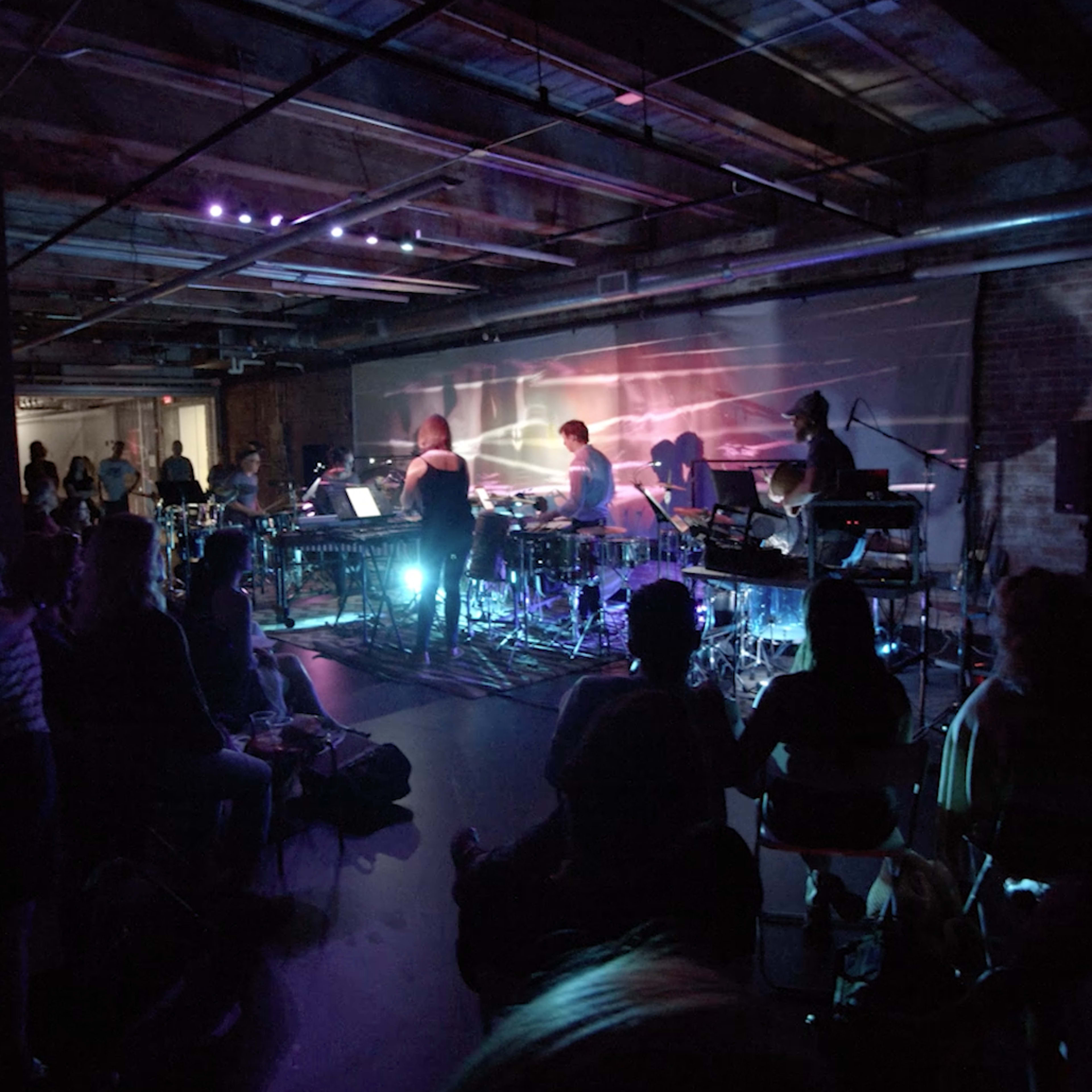 A crowd watches musicians perform onstage in a dimly lit venue with colorful lighting and various instruments.