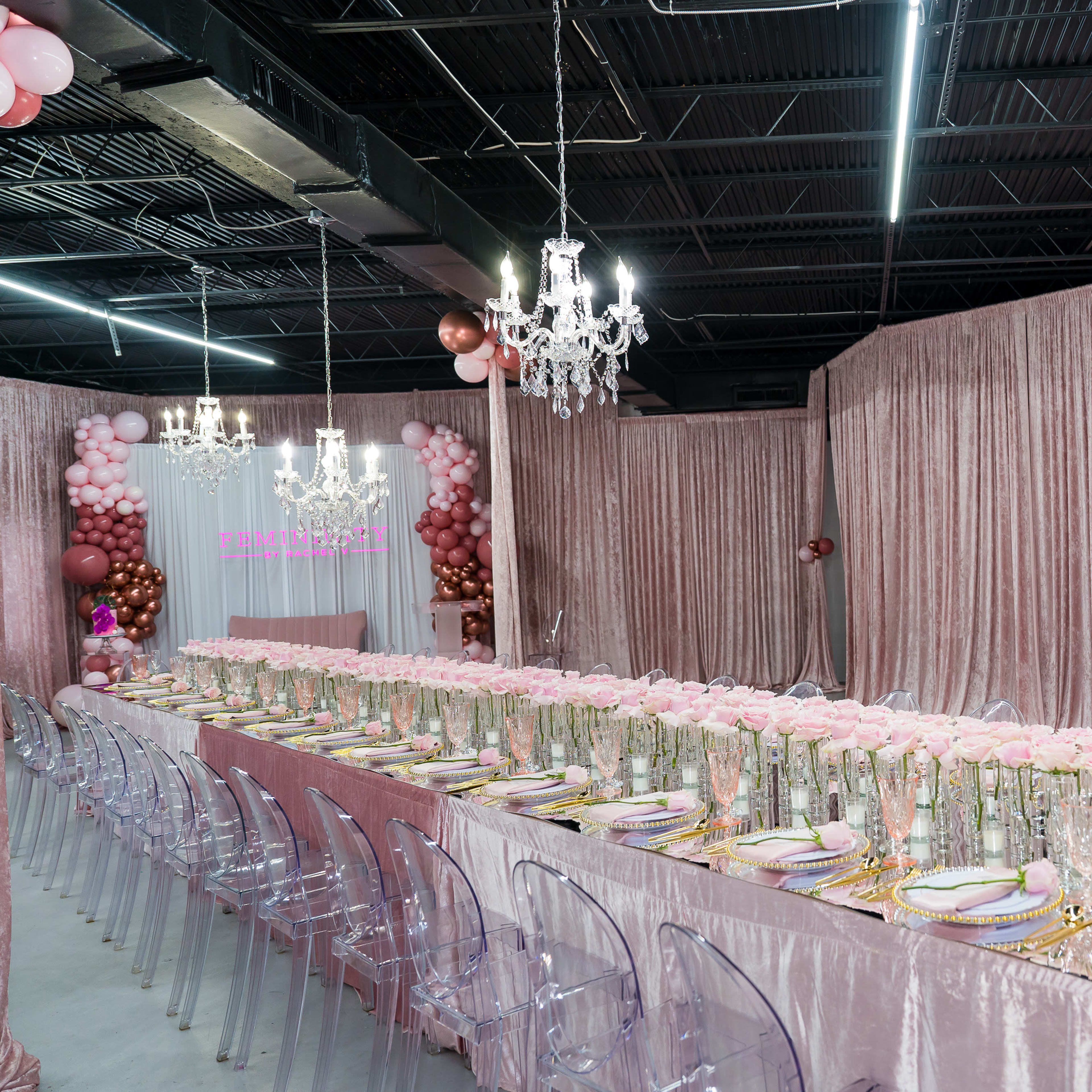A long table is set with pink tableware and floral centerpieces in a room adorned with draped fabric and decorative balloons.