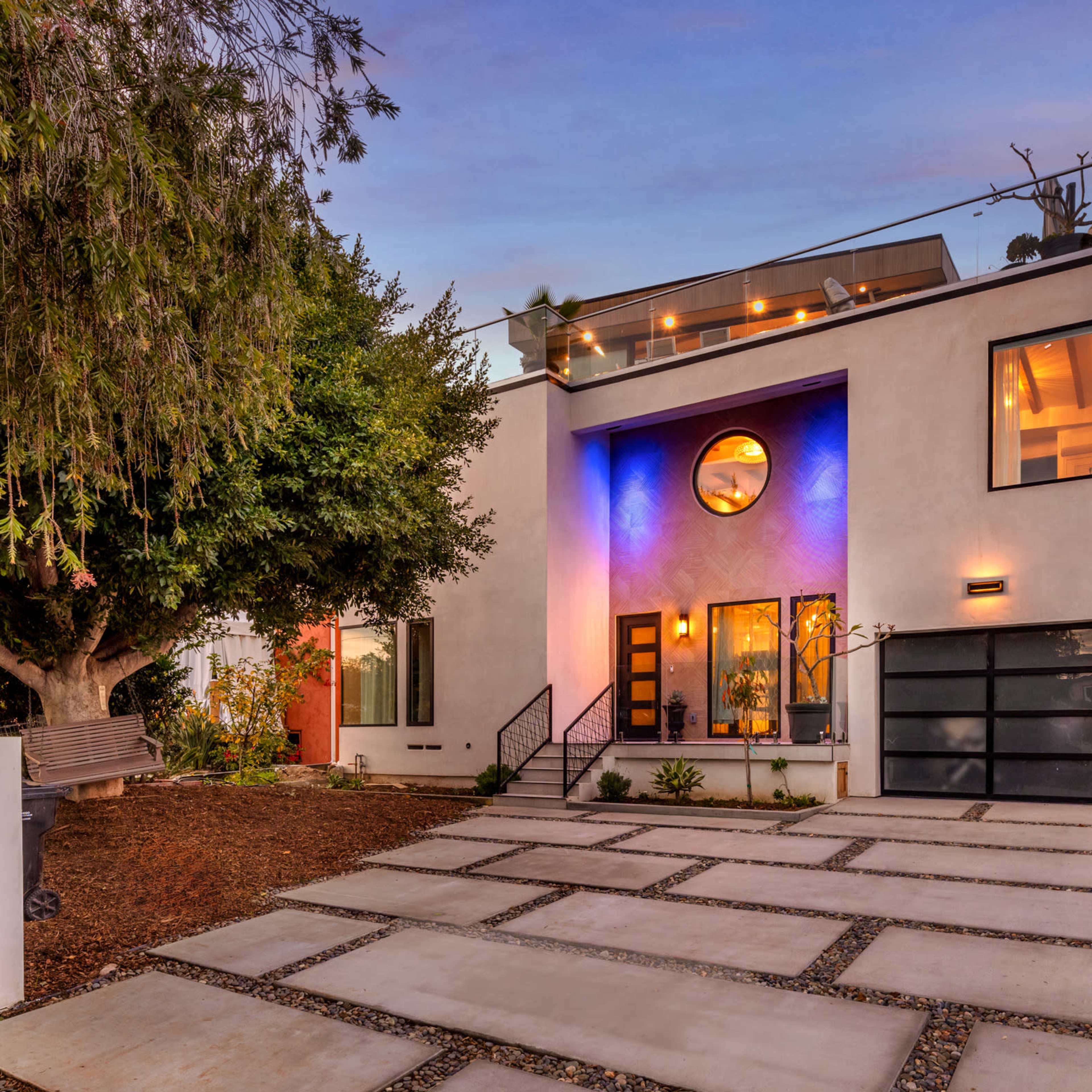 A modern two-story house features a curved window, a rooftop terrace, and a landscaped front yard with a stone walkway.