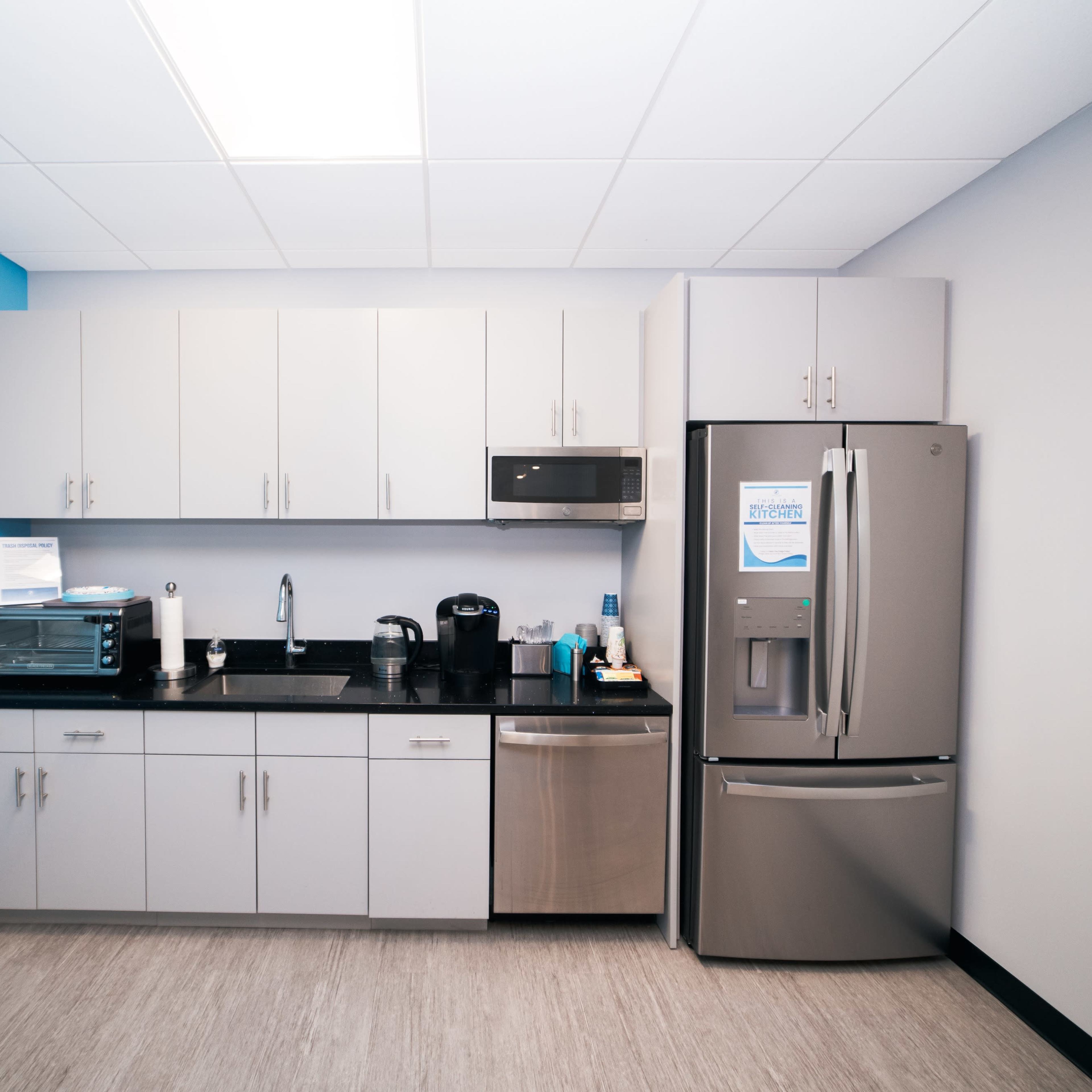 A kitchen area featuring gray cabinets, a stainless steel refrigerator, a microwave, and a coffee maker against a light blue wall.