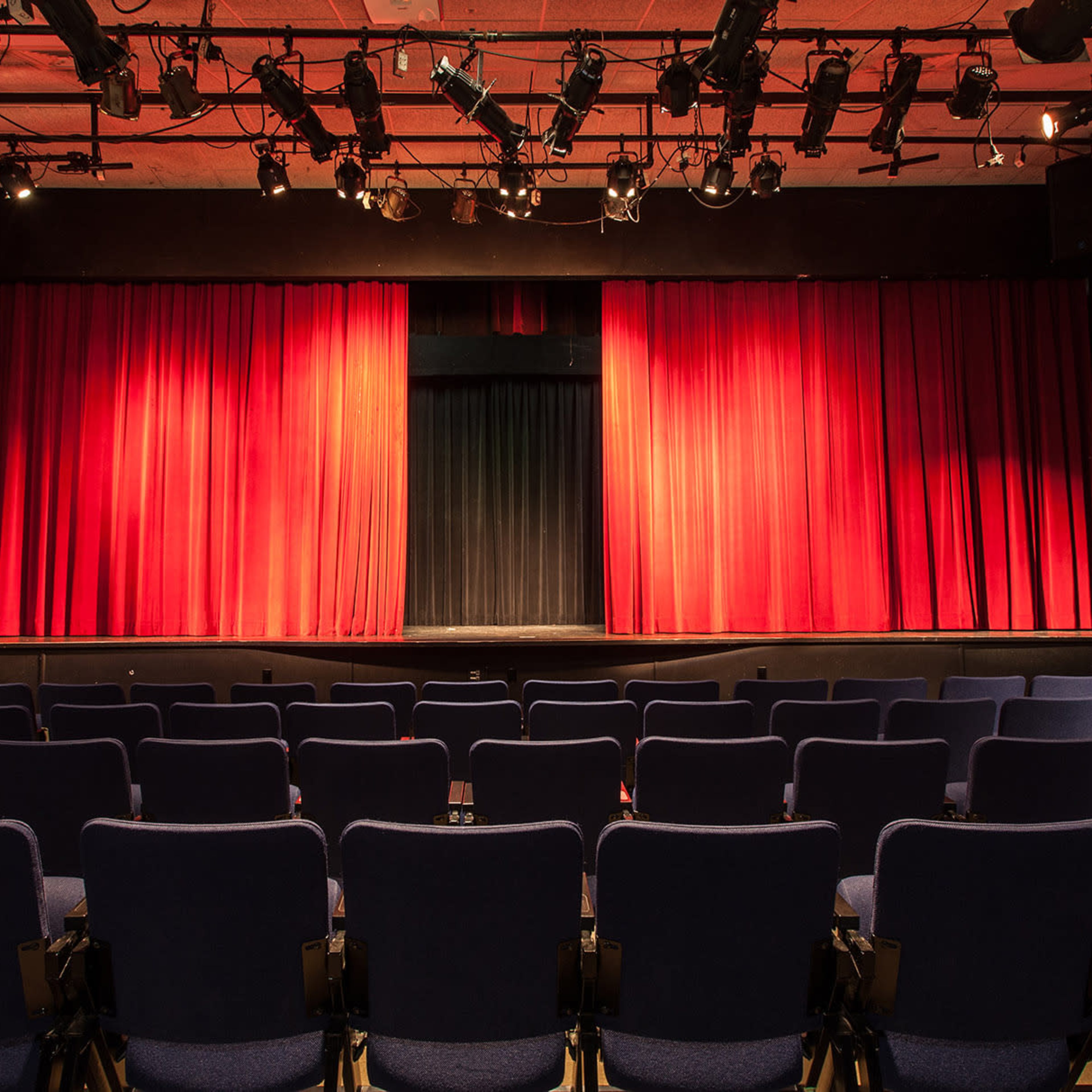 A theater stage with red curtains and a row of empty blue seats facing it.