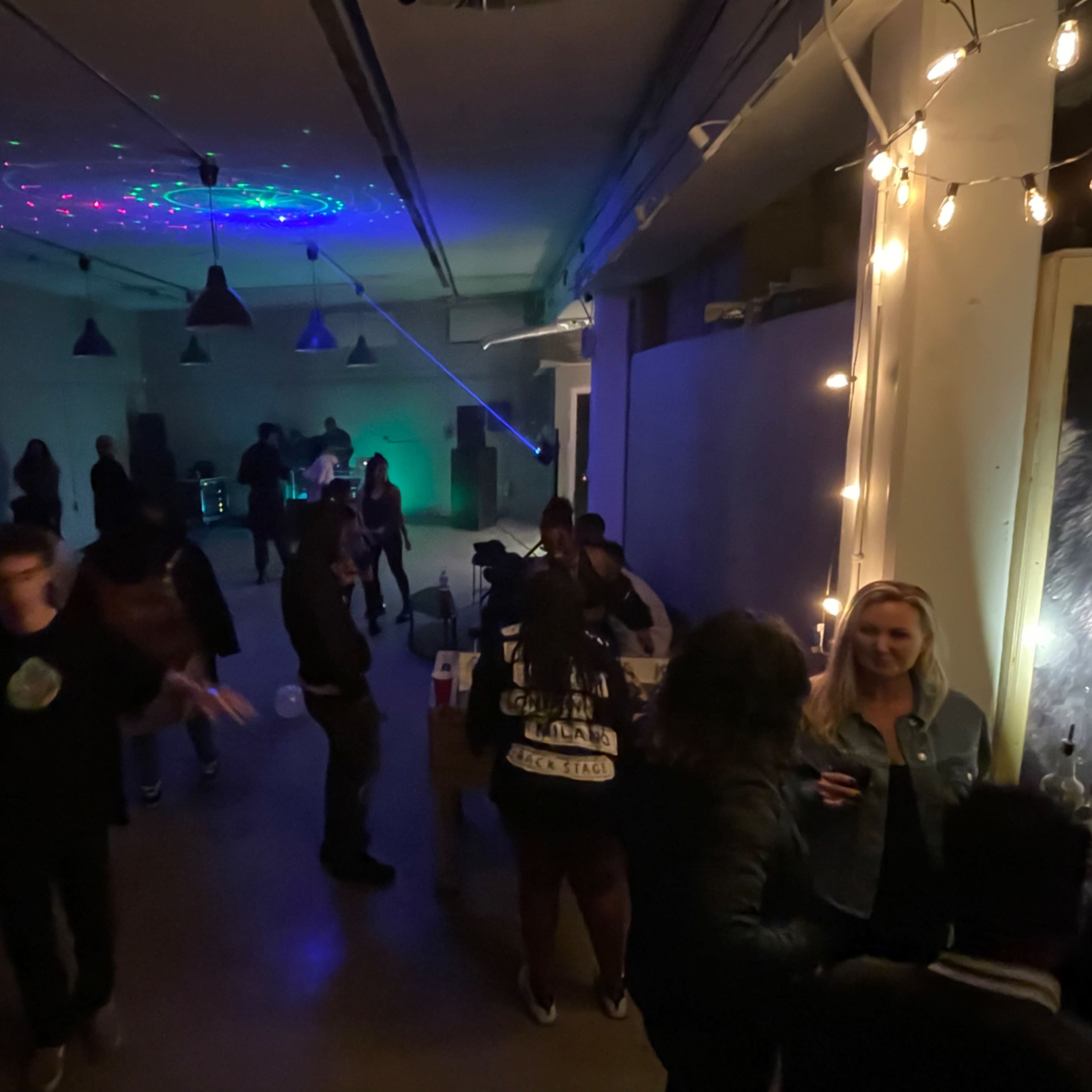 A group of people socializes in a dimly lit room with colorful lights and a bar setup.