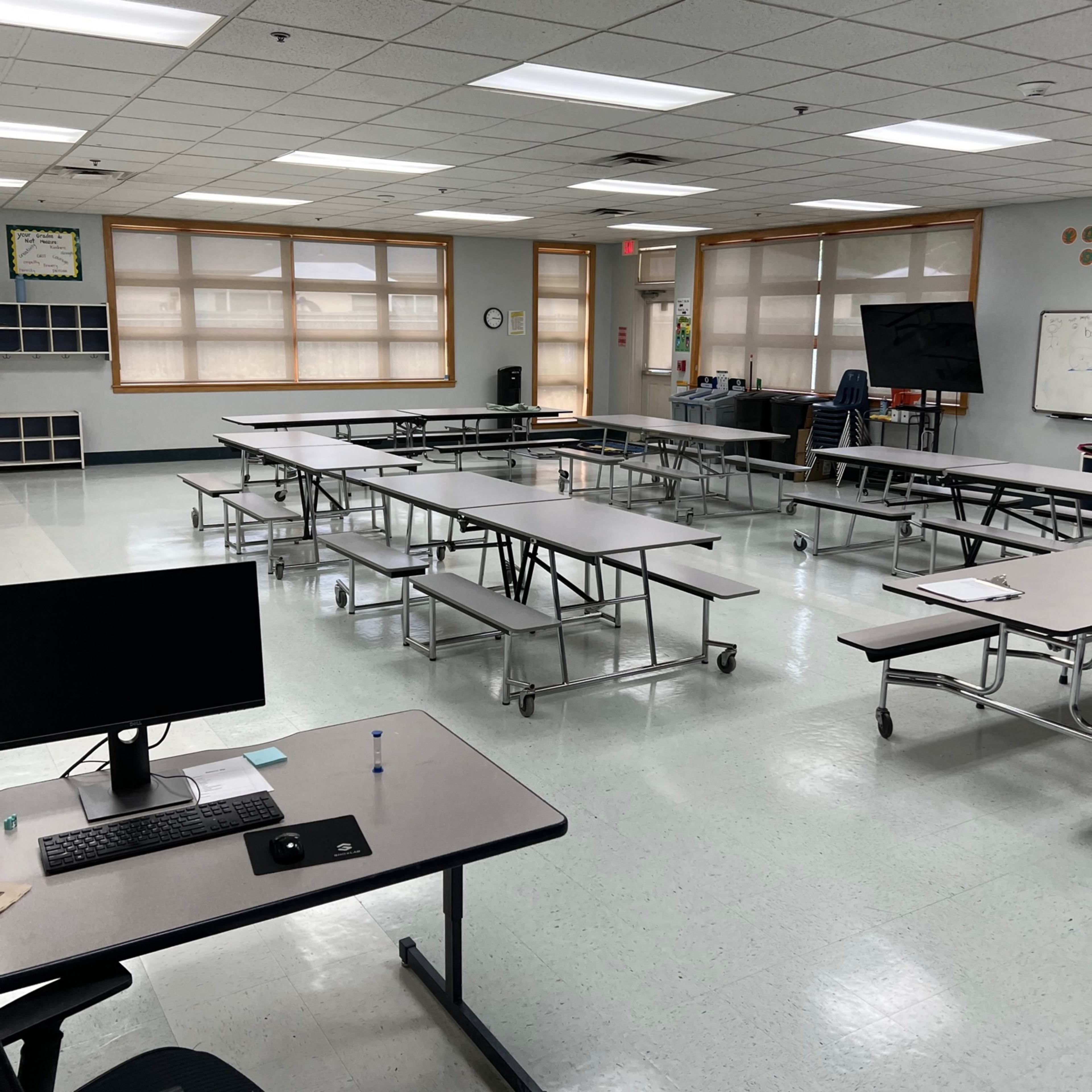 An empty classroom with several tables arranged in rows, a computer on a desk, and large windows allowing natural light.