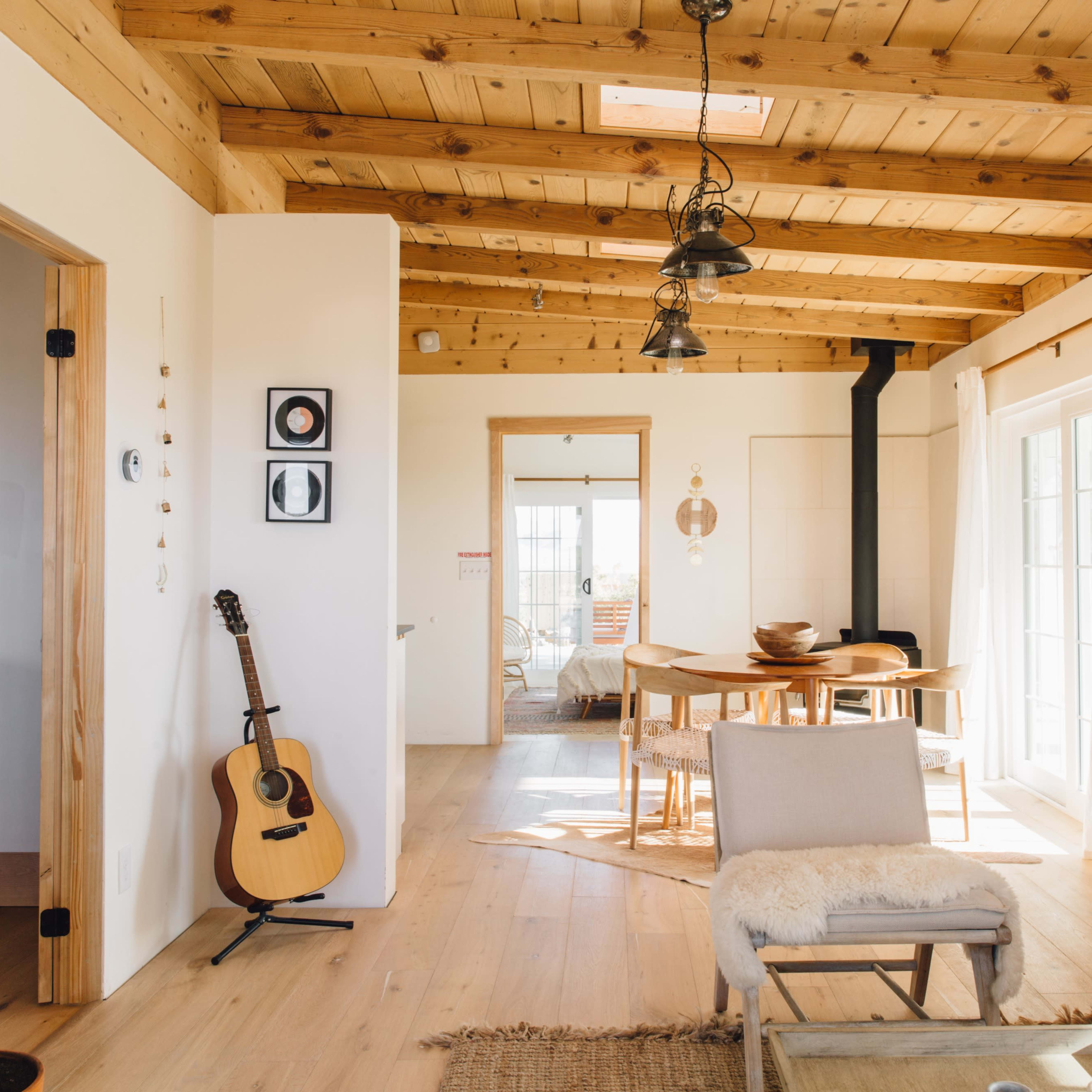 A bright, open living space with wooden beams, a guitar leaning against the wall, and a round dining table surrounded by chairs.