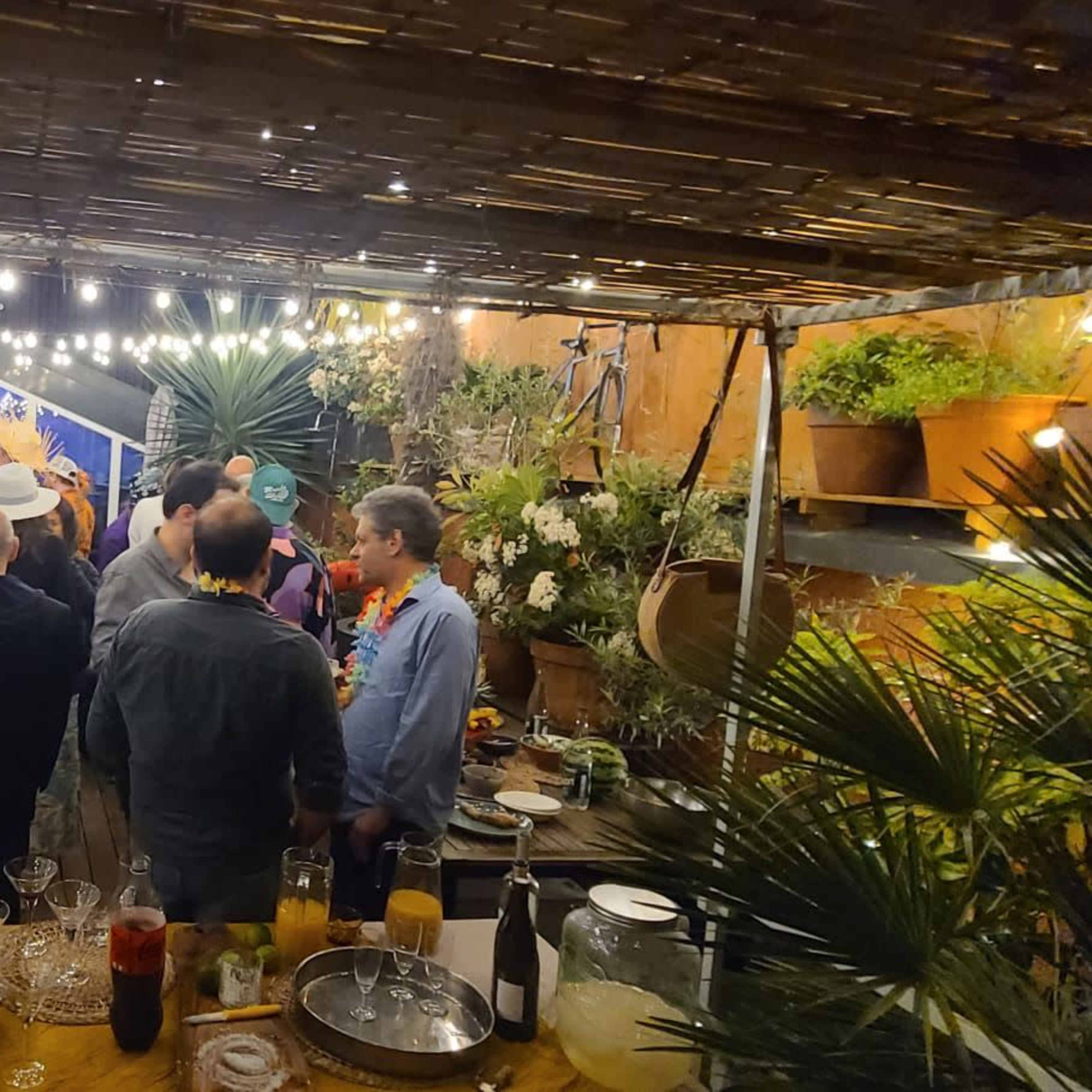 A group of people gathers at a decorated outdoor space, surrounded by plants and soft lighting, while enjoying drinks and conversation.