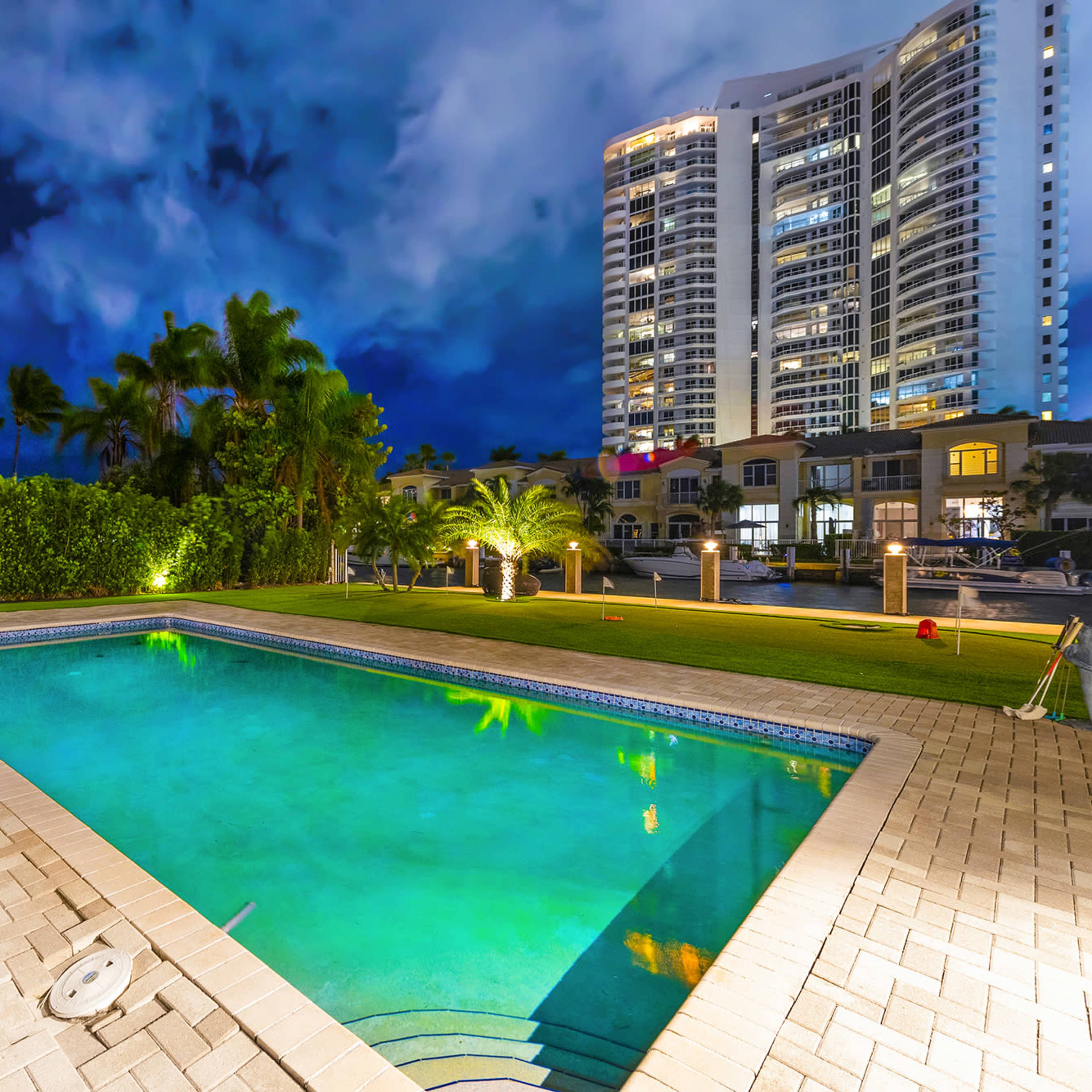 A well-lit swimming pool surrounded by palm trees and modern buildings under a dramatic night sky.