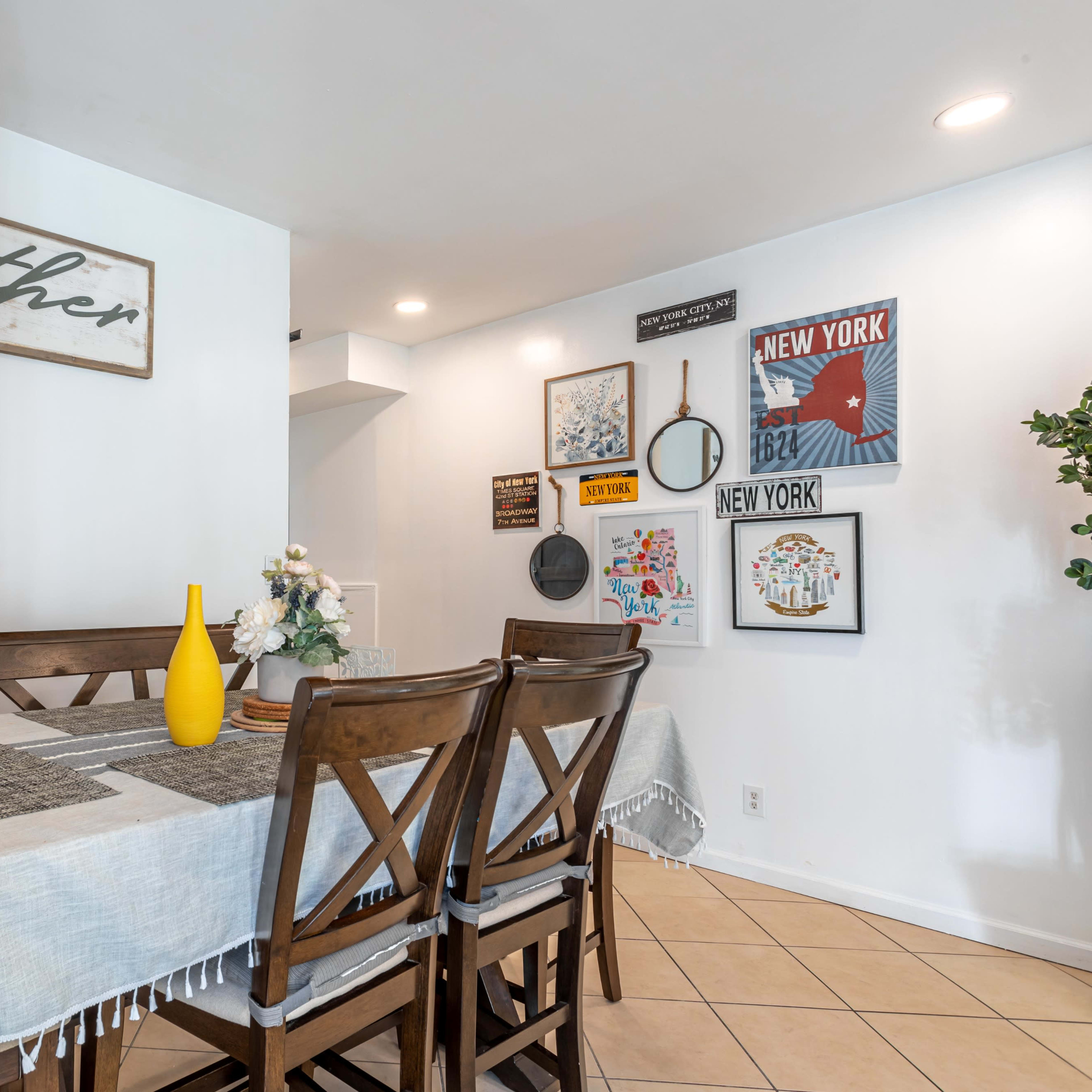 A dining area features a table set with a light-colored tablecloth, wooden chairs, a yellow vase, and wall decorations including signs and framed artwork.