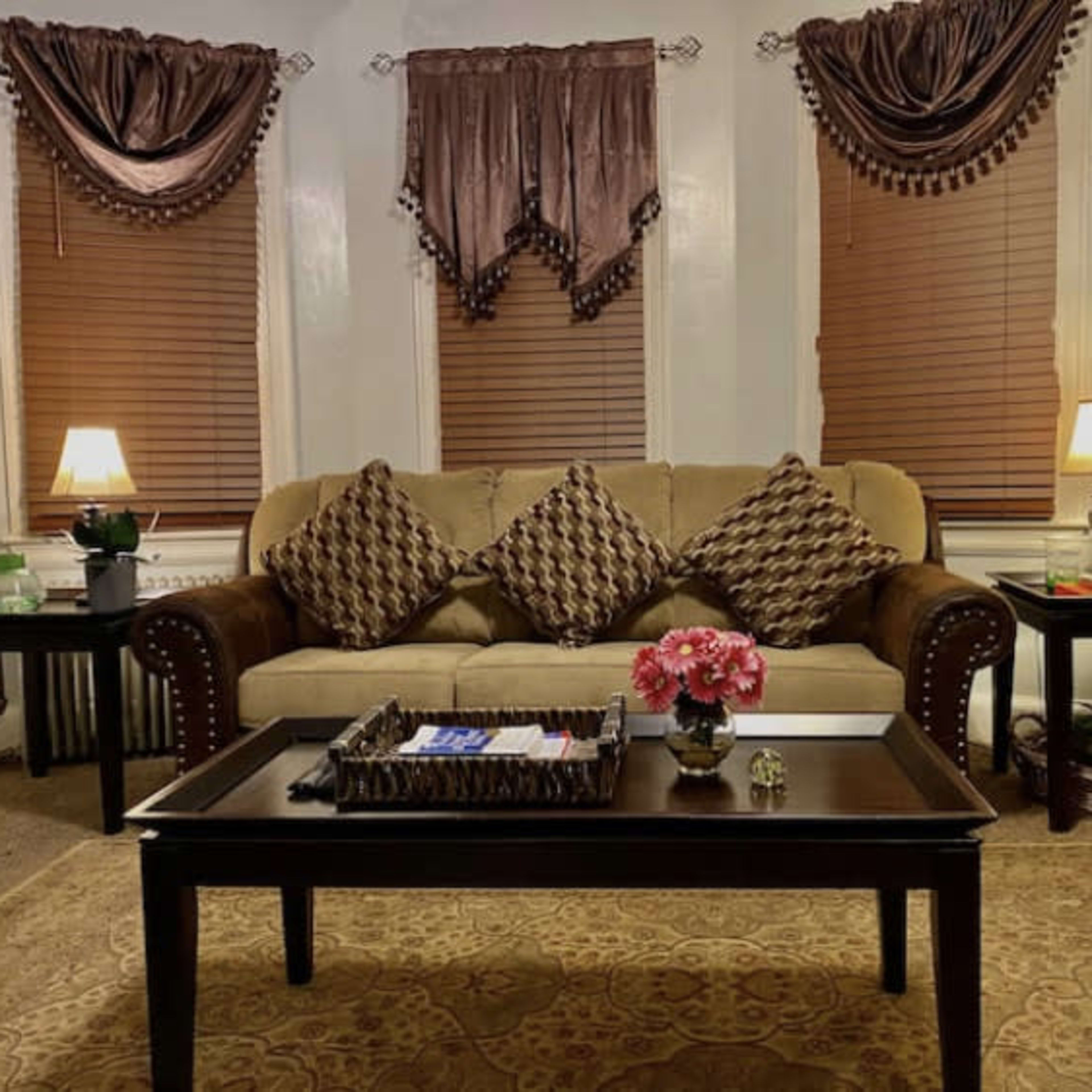 The scene depicts a living room with a beige sofa adorned with decorative cushions, accented by two tables with lamps and plants, all set against a backdrop of brown curtains and a patterned rug.
