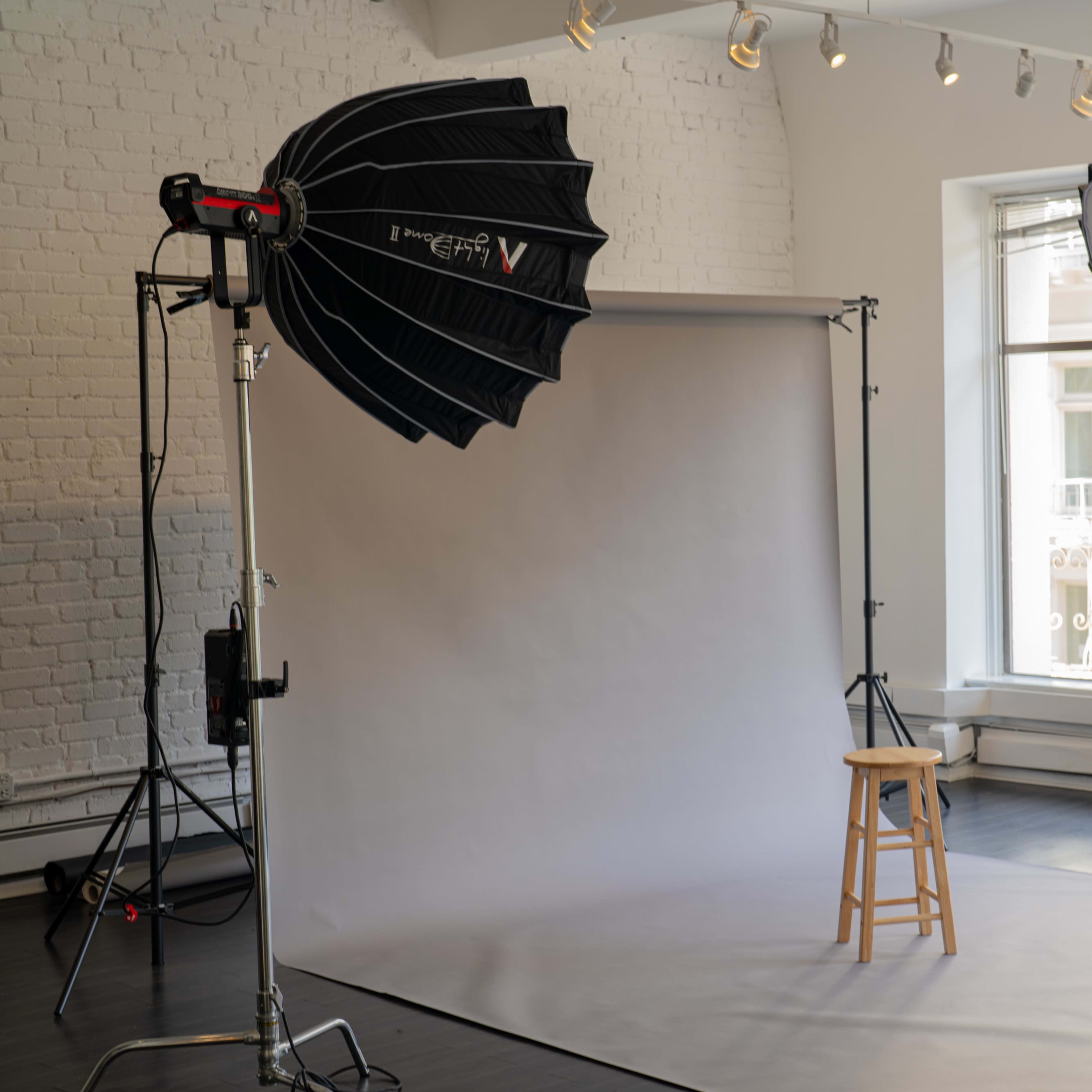 A photography studio setup with two softbox lights and a blank backdrop, featuring a stool positioned in front of it.