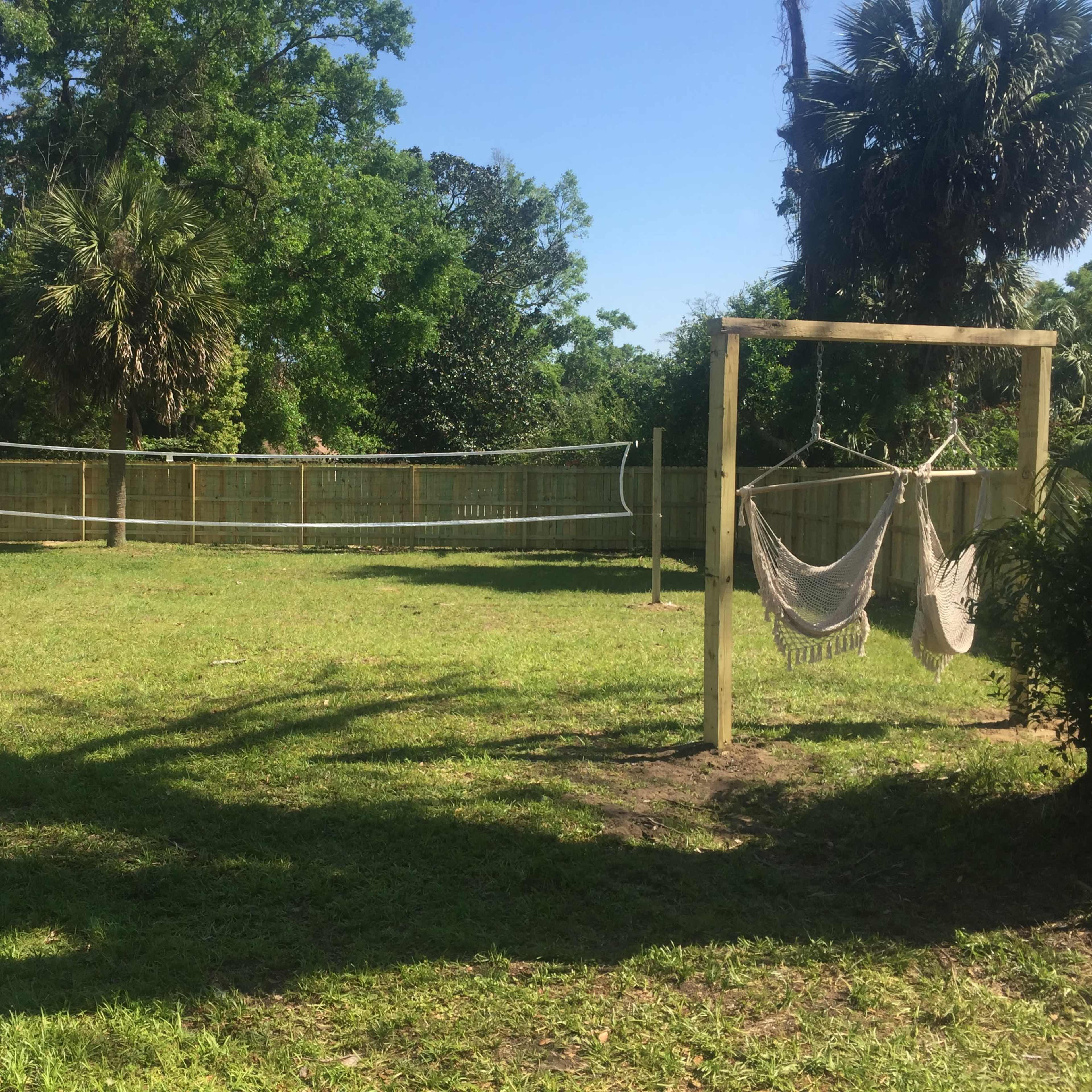 A grassy area enclosed by a wooden fence, featuring a volleyball net and a hammock stand.