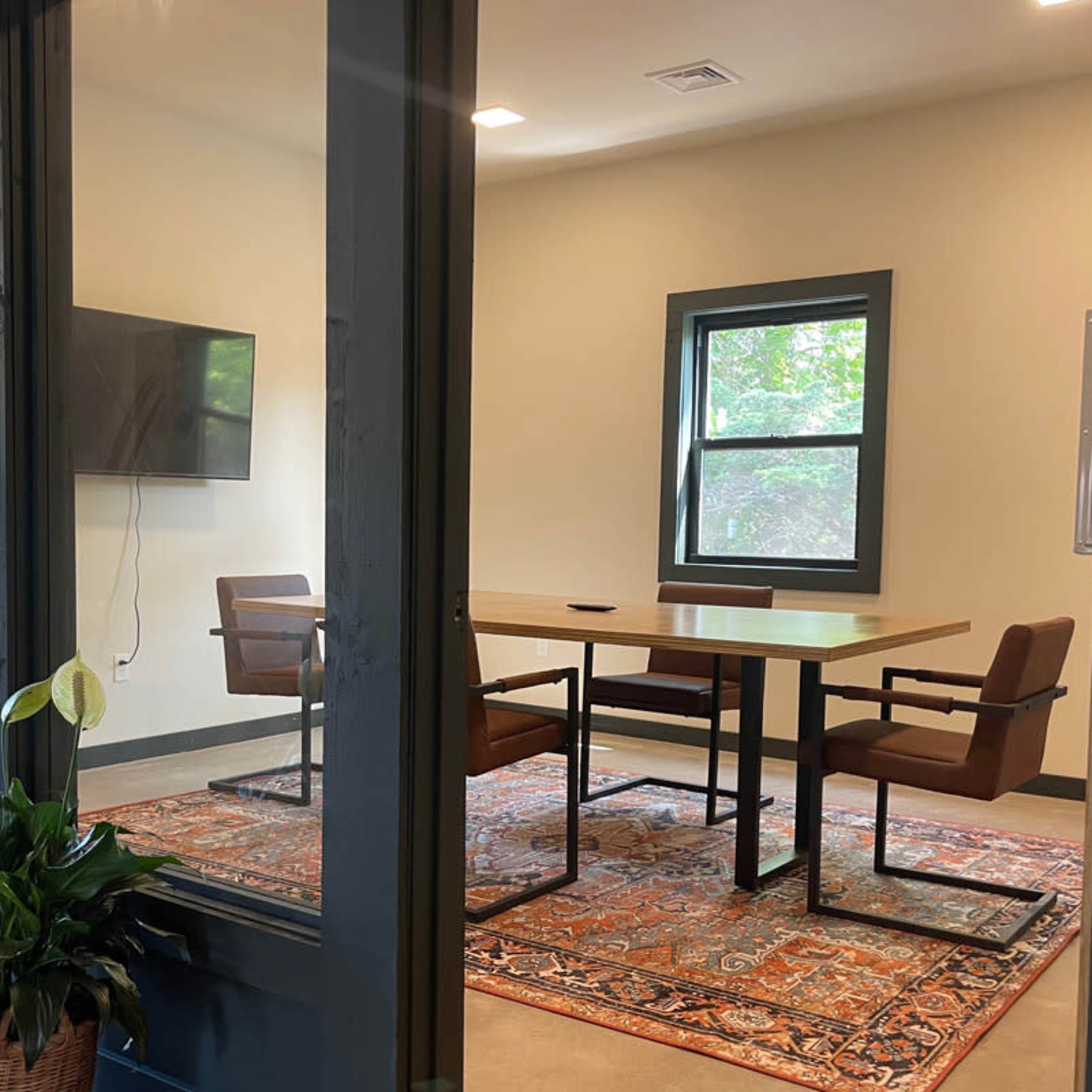 A meeting room features a table with four chairs, a large television on the wall, and a decorative rug, framed by glass doors and a potted plant nearby.