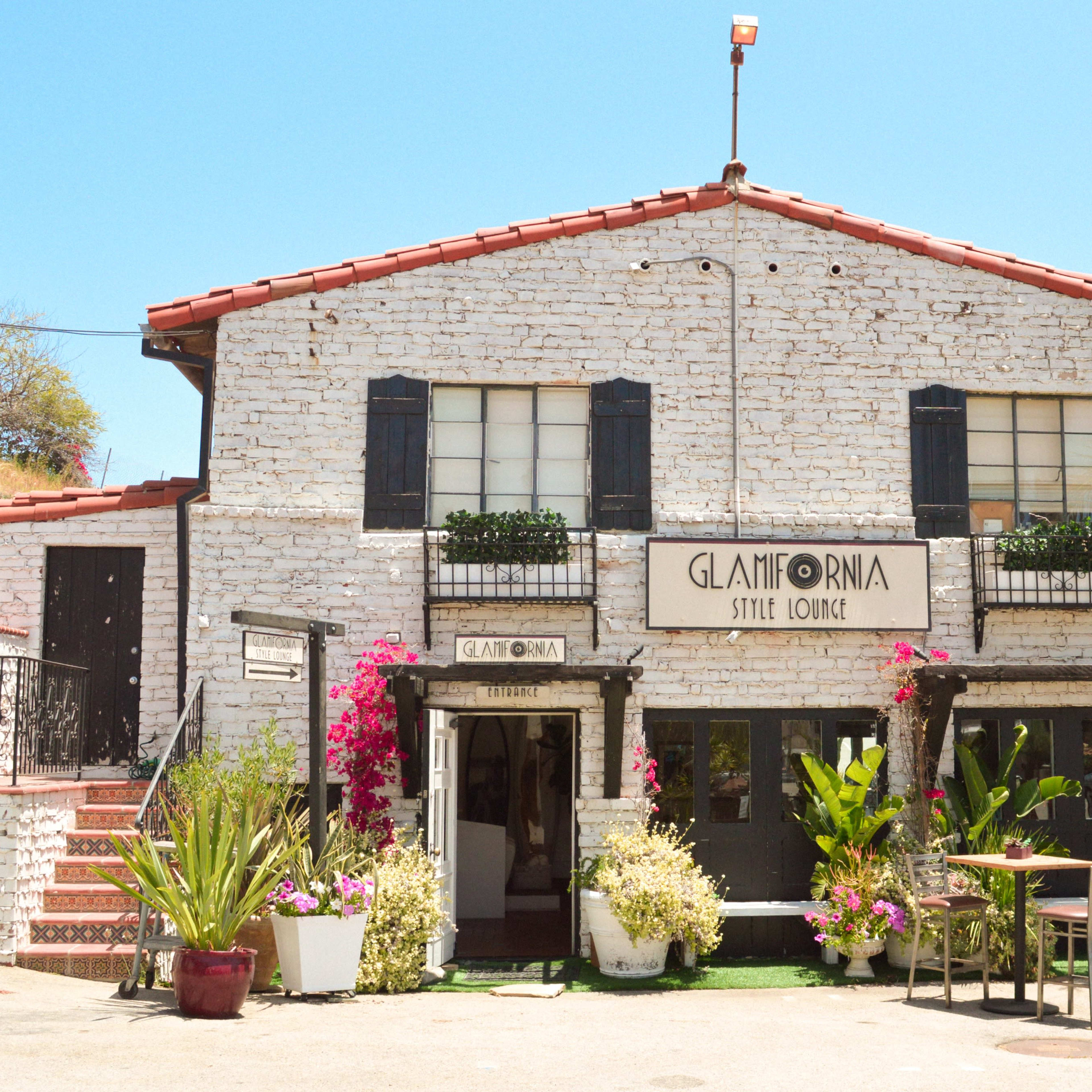 A two-story building with a white brick facade, black shutters, and a sign reading "Glamifornia Style Lounge," surrounded by colorful plants and palm trees.