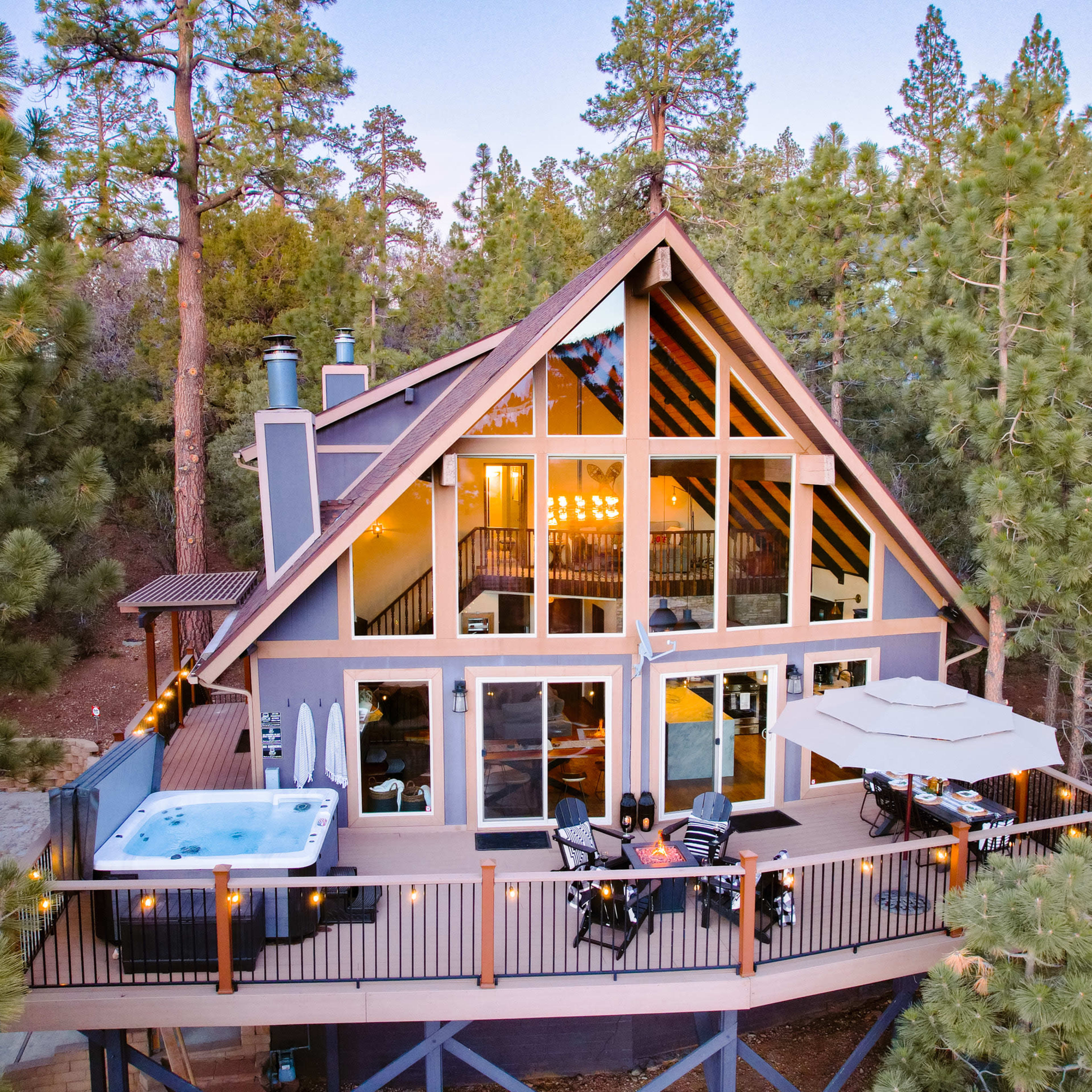 A modern A-frame cabin surrounded by tall pine trees features large windows, a deck with outdoor seating, and a hot tub.