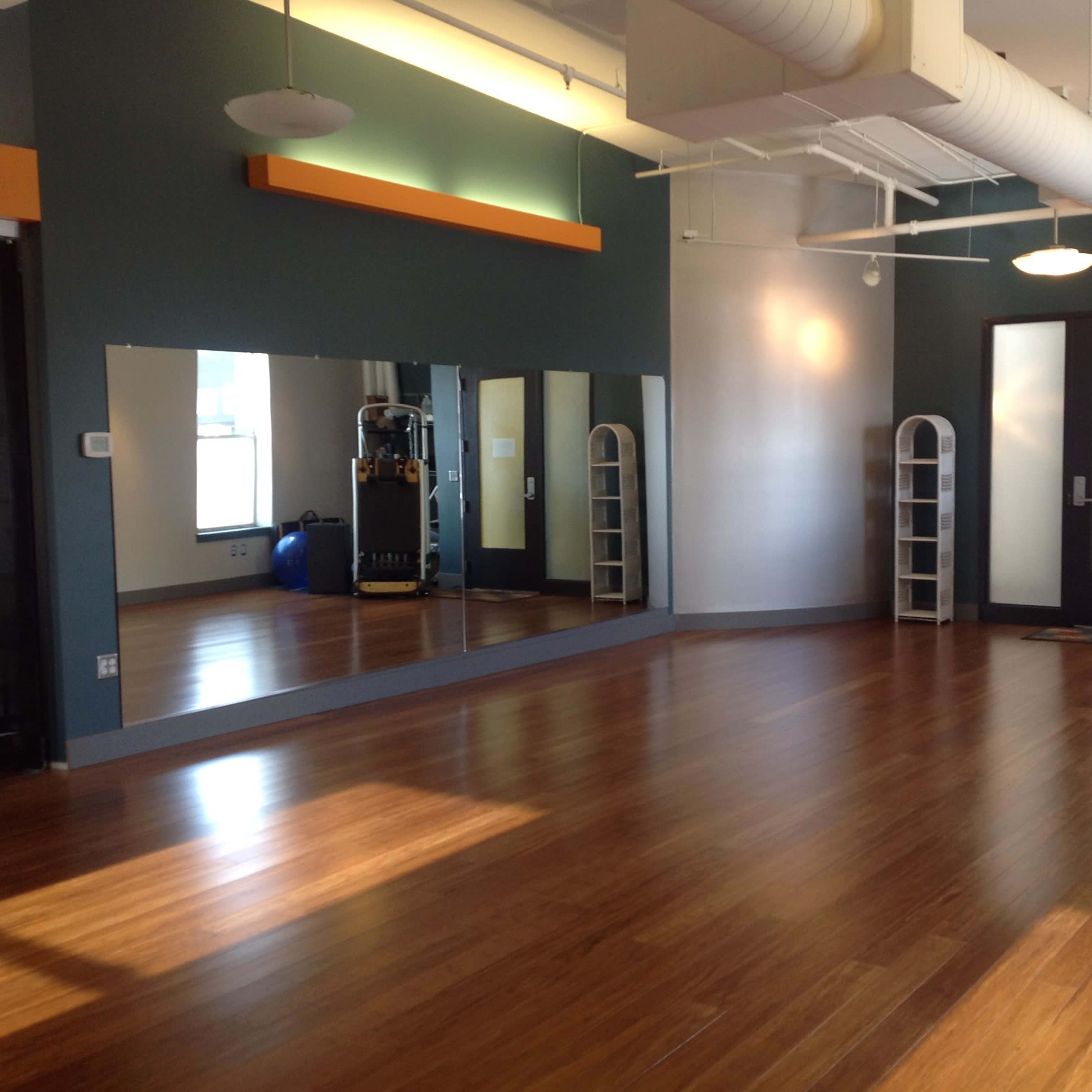 An empty fitness studio with hardwood floors, a large mirror along one wall, and exercise equipment in the corner.