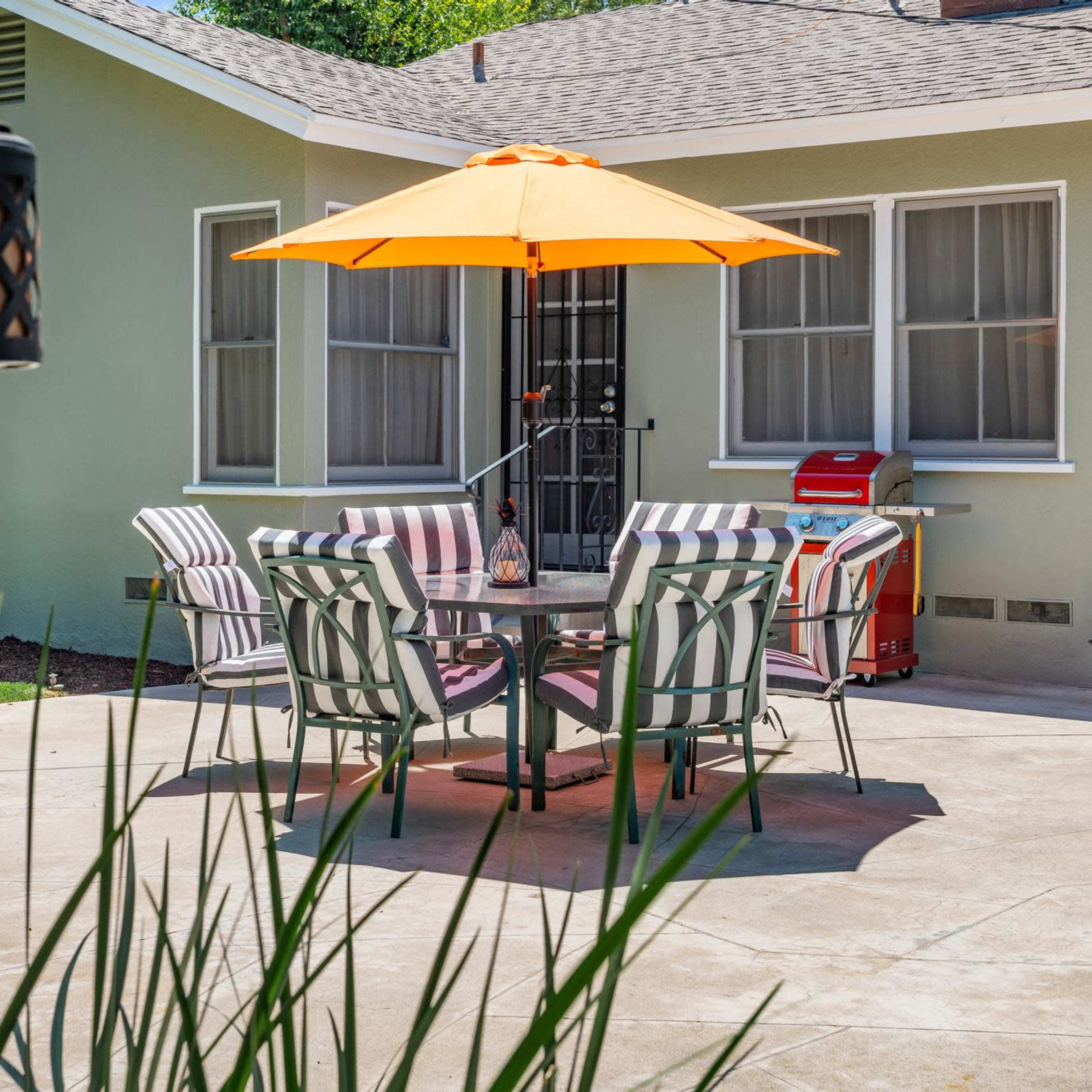 A patio features a round table with striped chairs and an orange umbrella, surrounded by a green lawn and a light-colored house in the background.
