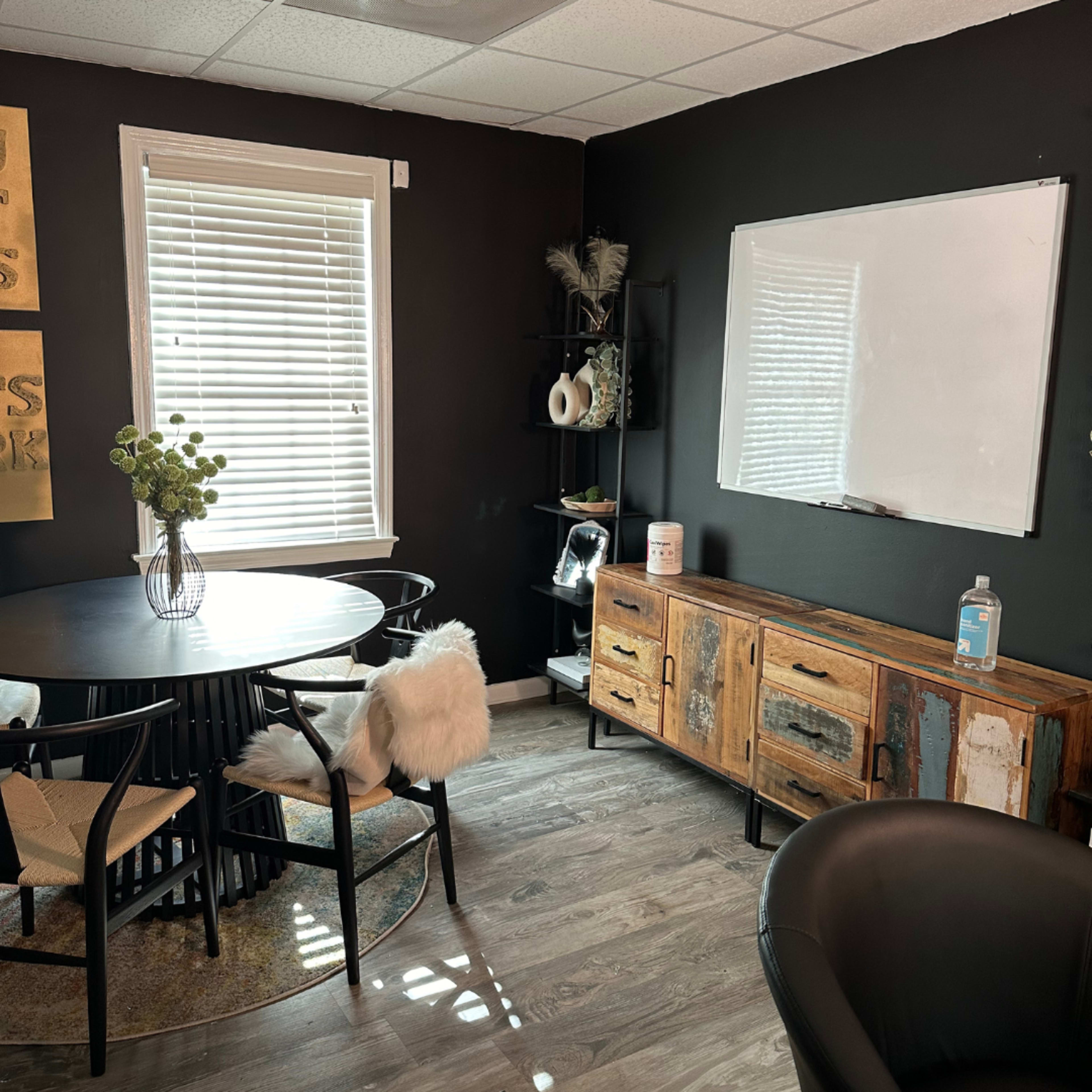 A modern interior office space featuring a round dining table, an antique wooden sideboard, a whiteboard, and a bookshelf with decorative items against a dark wall.