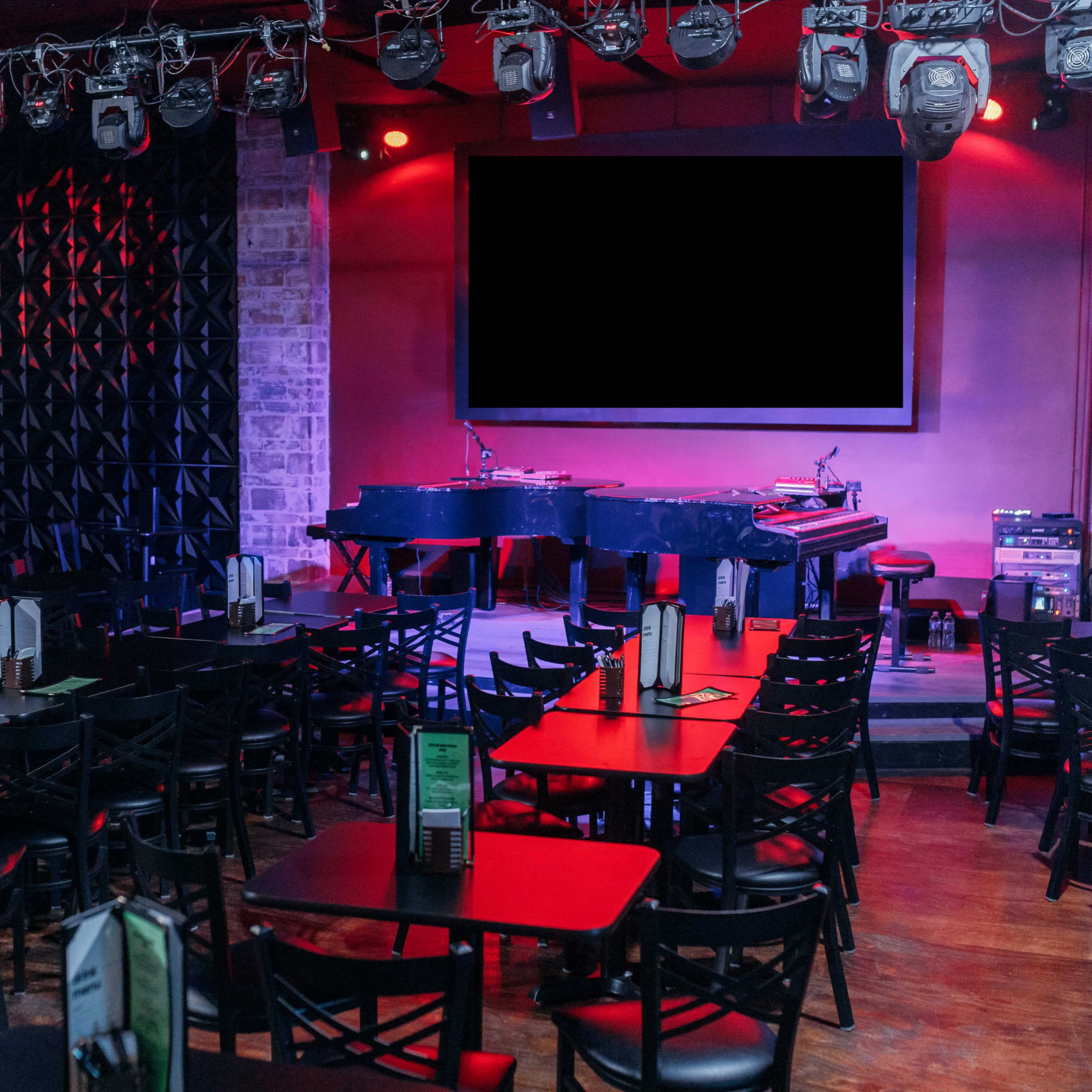 An empty performance venue with rows of black tables and chairs facing a small stage that features a grand piano and a large screen.
