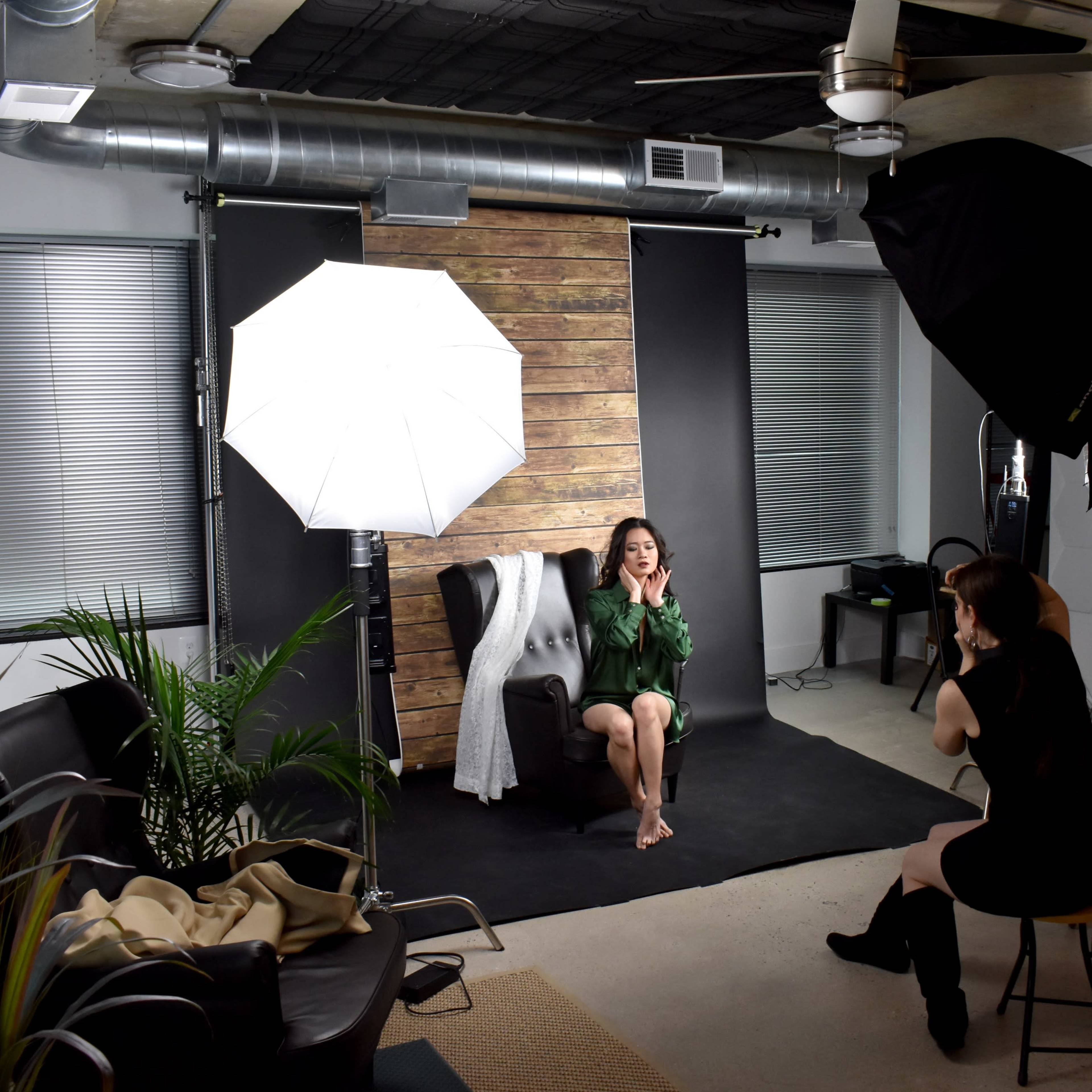 A model sits on a chair in a photography studio, surrounded by soft lighting equipment and backdrops, while another person adjusts the scene.