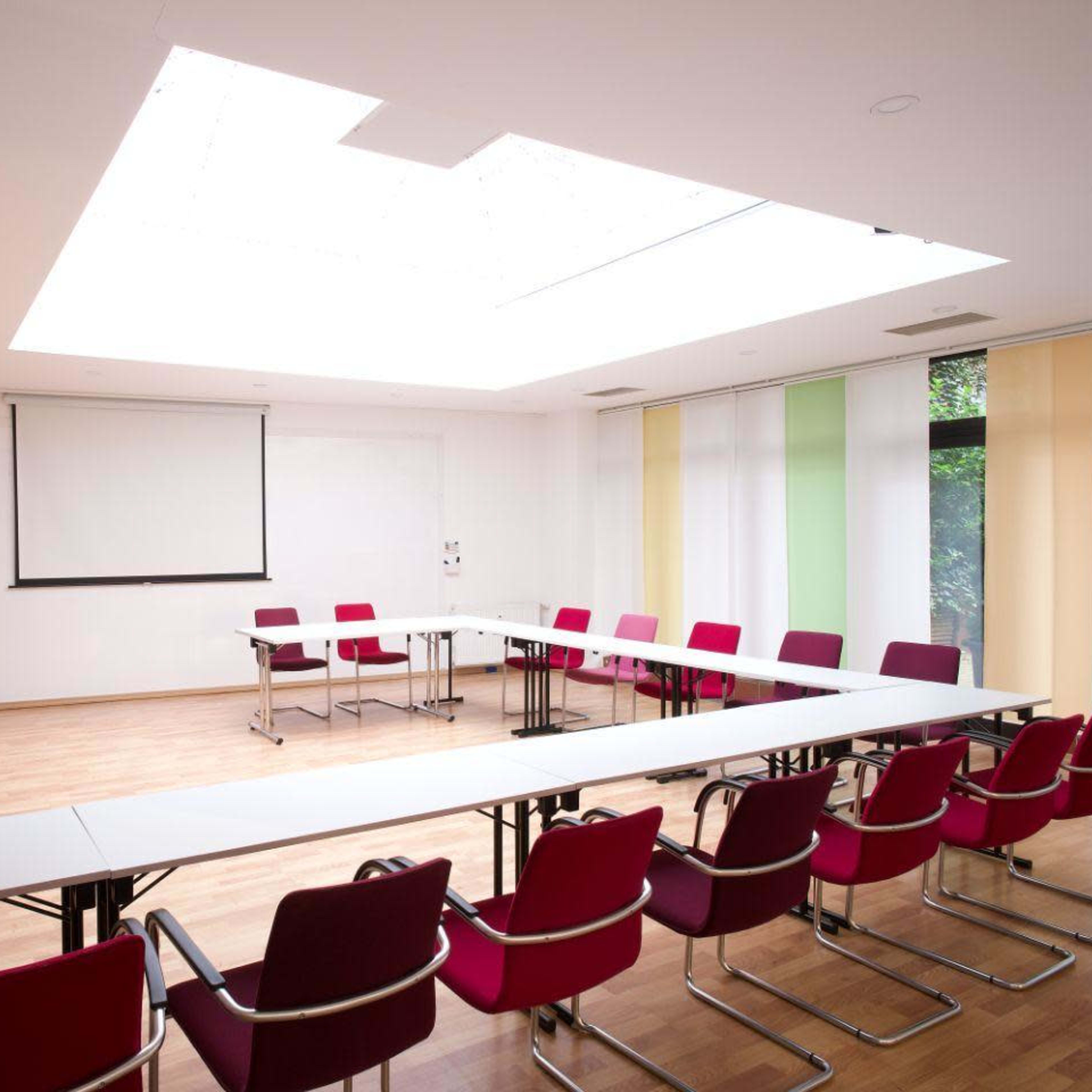 A conference room features long tables arranged in a U-shape with red chairs, illuminated by a skylight above.