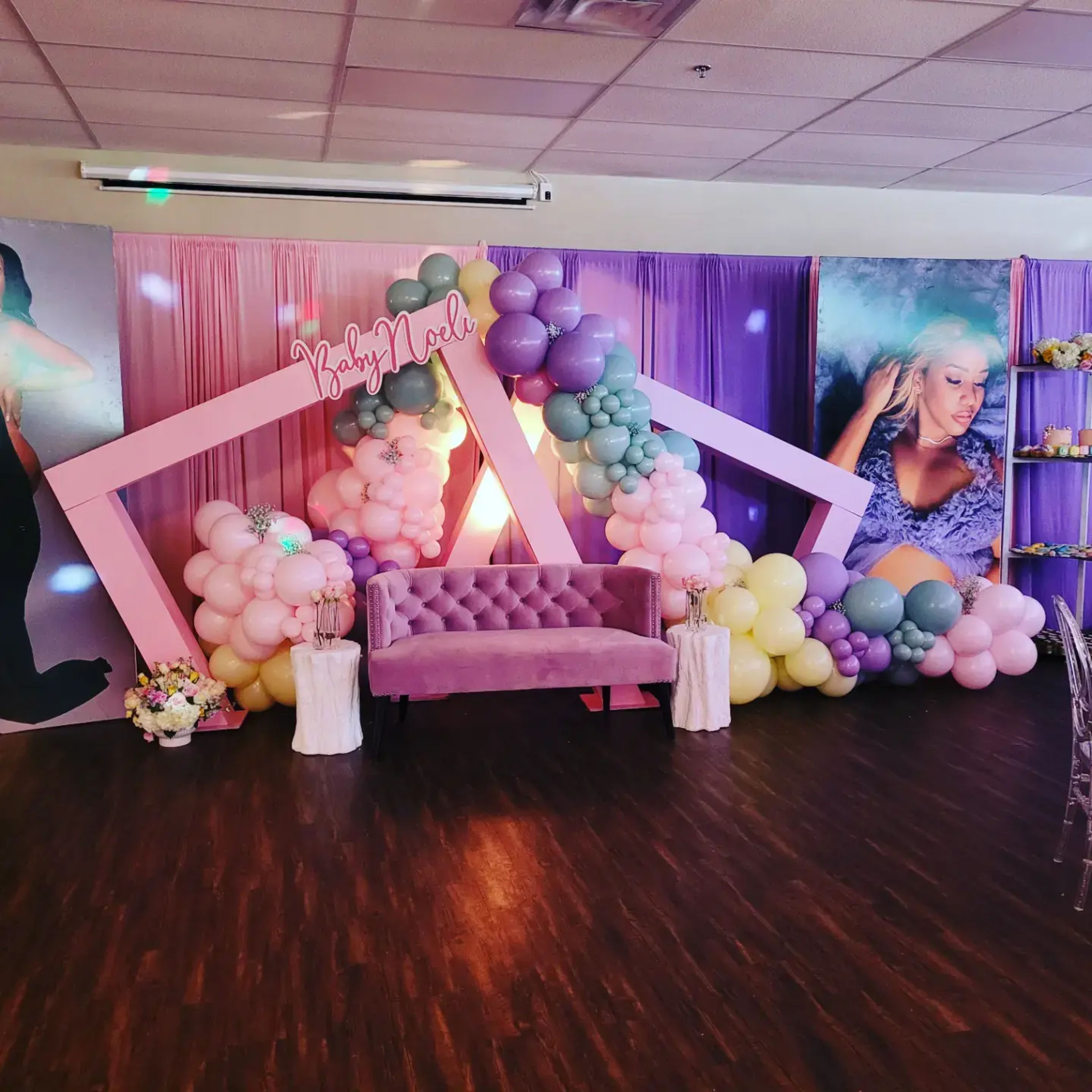 A decorated event space featuring a pink couch surrounded by a balloon arch and a backdrop with large images of a woman.