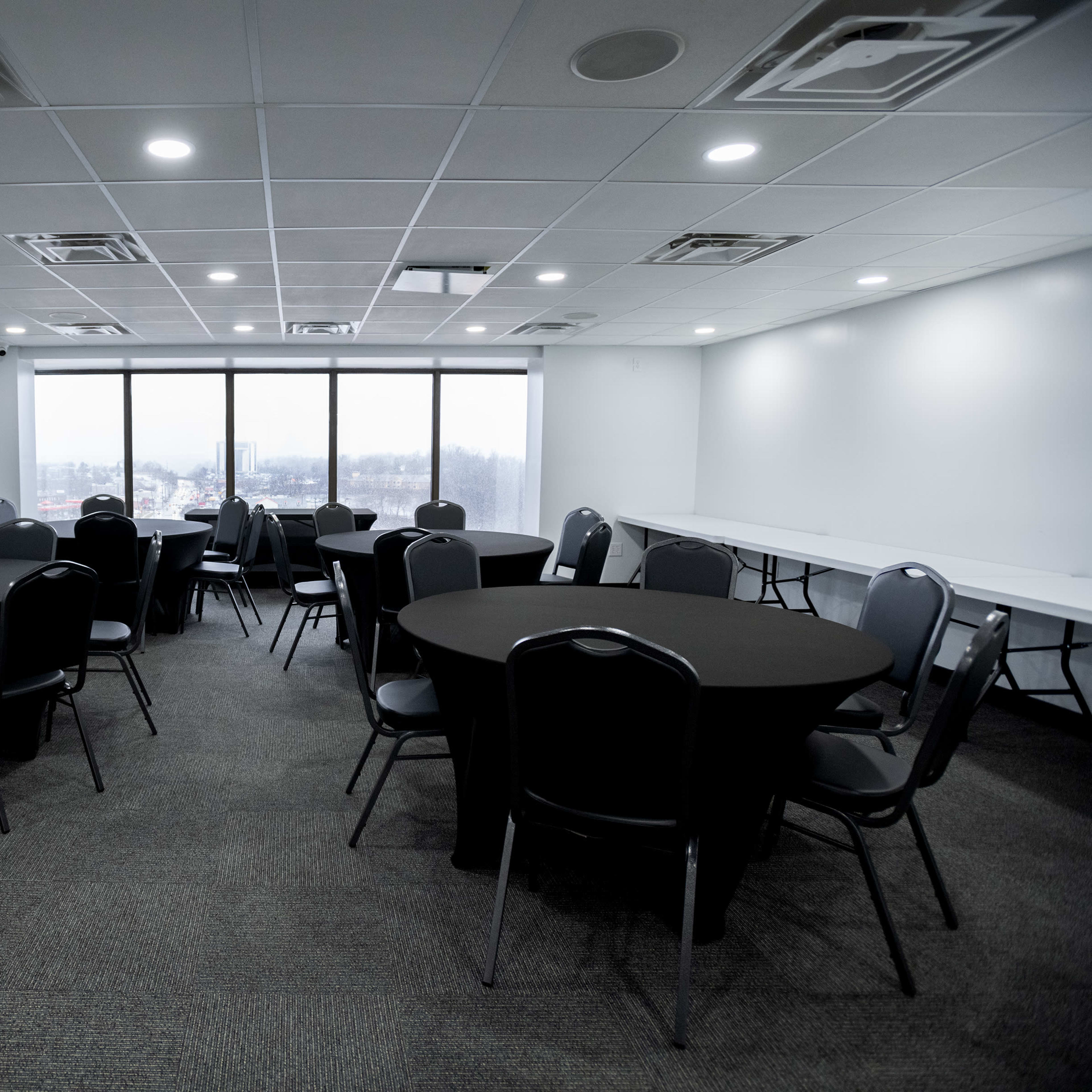 A spacious room with several round tables covered in black linens and a row of windows allowing natural light.