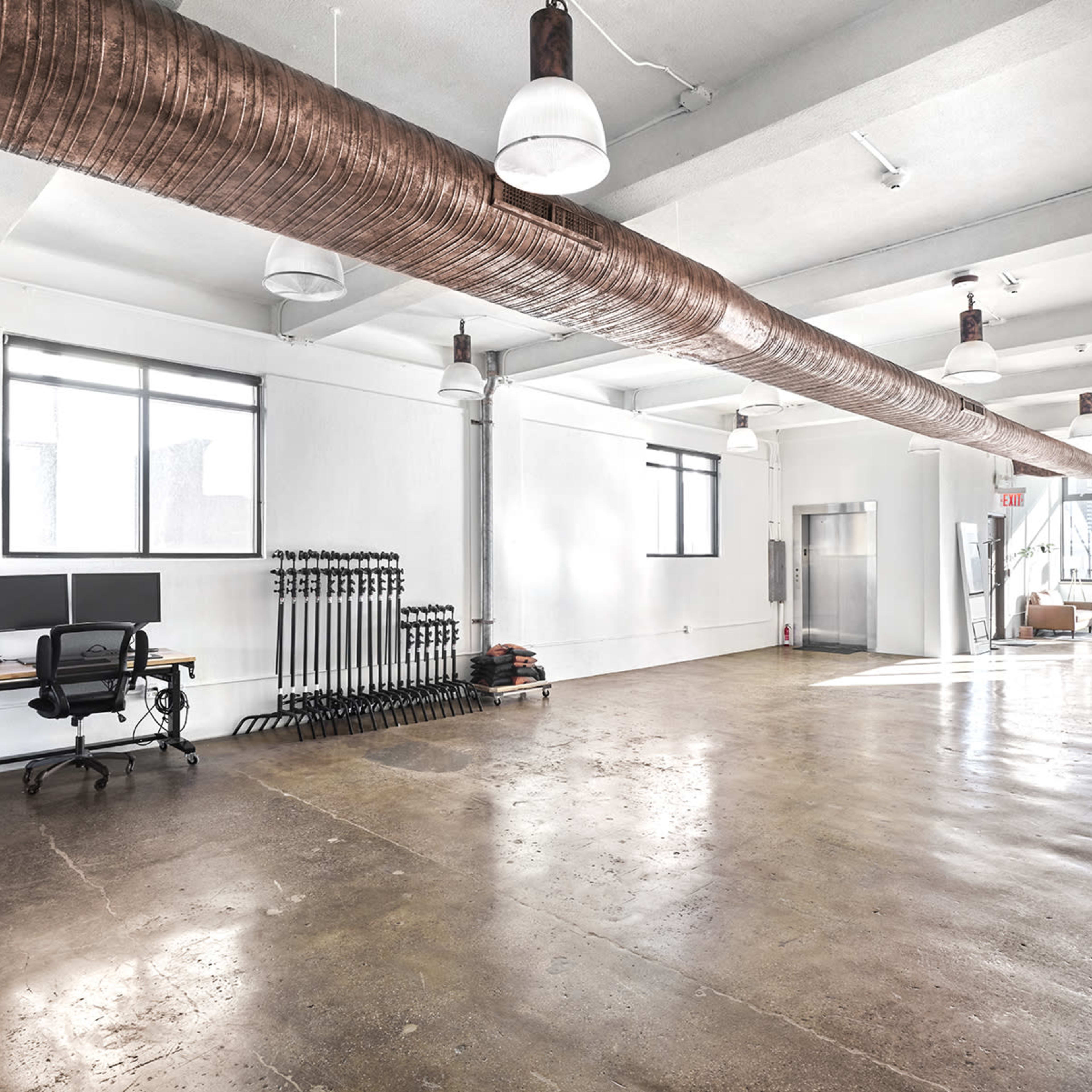 A spacious, empty room with concrete flooring, large windows, and a workstation featuring two computer monitors.