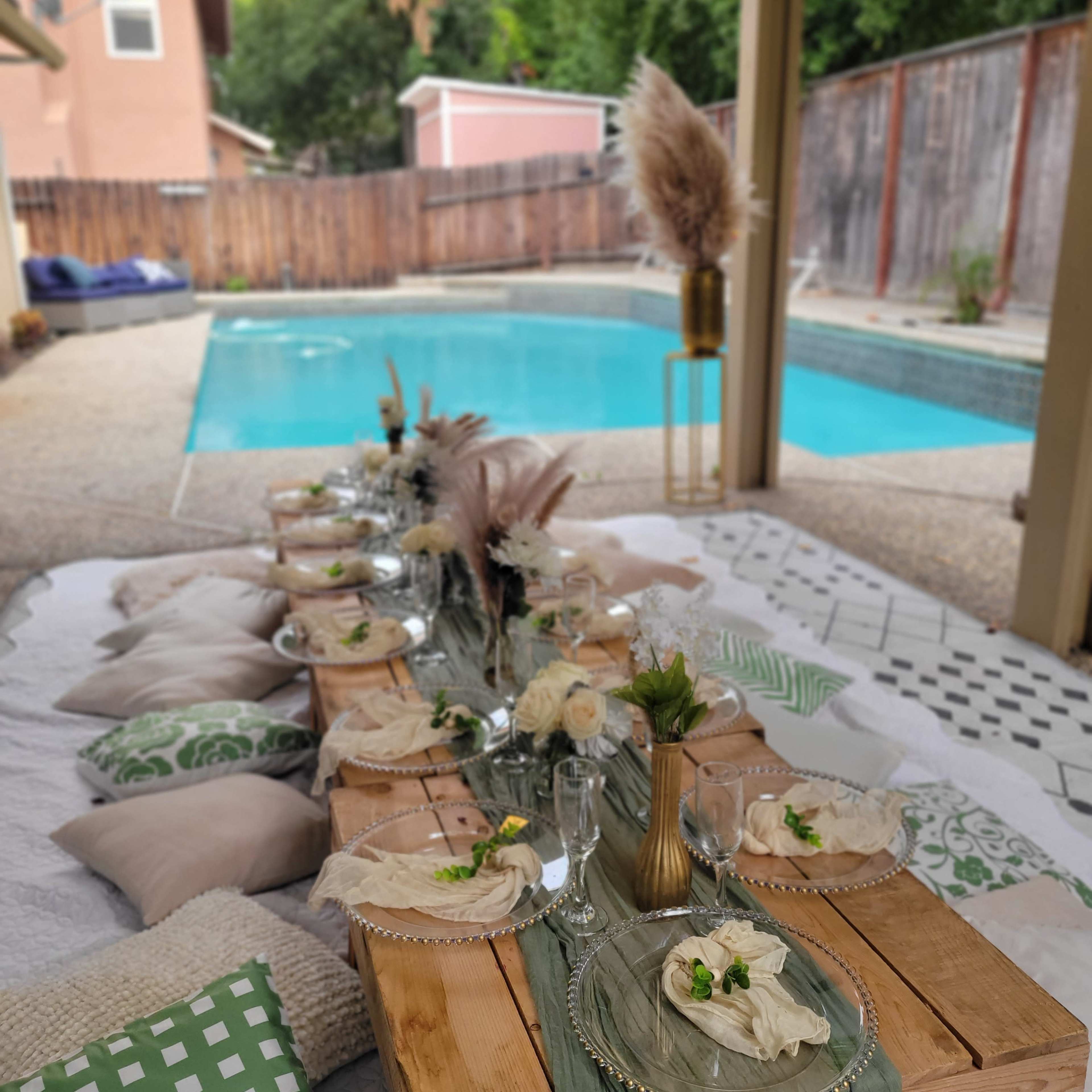 A long wooden table is set with dishes and glassware, surrounded by decorative cushions, near a swimming pool under a covered patio.